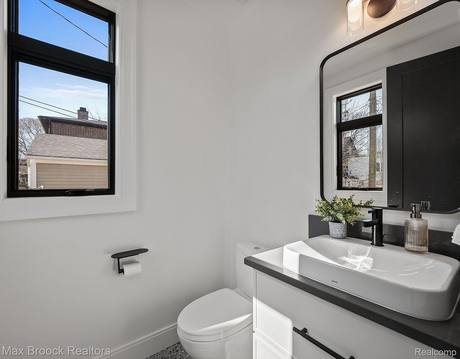 This is a well-lit guest bathroom featuring a modern vanity with a white vessel sink and black hardware. A sleek black-framed mirror hangs above the sink, reflecting a window. The toilet is white, and the walls are painted a clean white, creating a bright and inviting space.