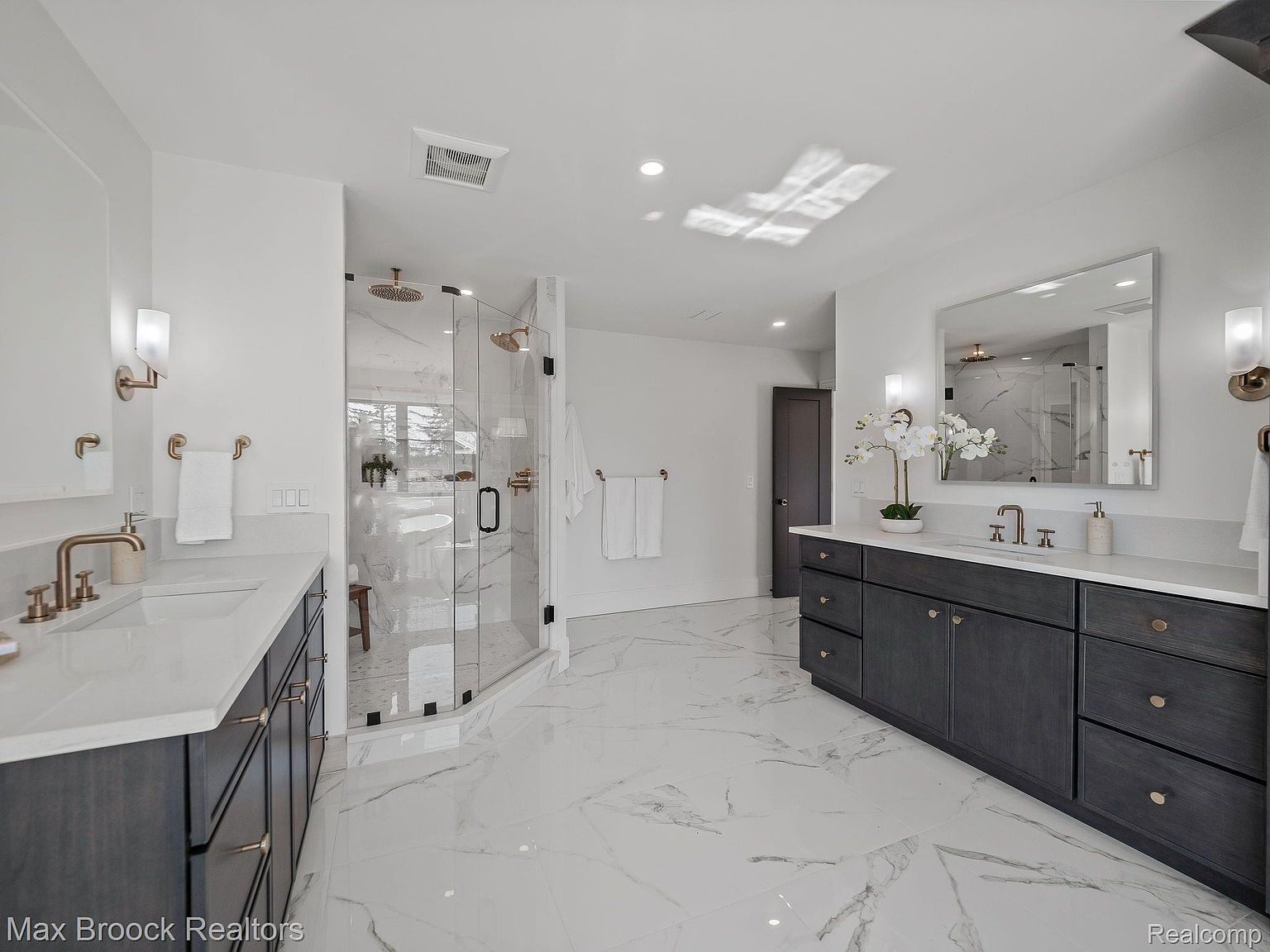 This is a luxurious primary bathroom featuring a double vanity with dark wood cabinets and gold hardware, complemented by a large mirror and modern sconces. The bathroom boasts a spacious glass-enclosed shower with dual shower heads and marble-like tile flooring, creating a bright and elegant atmosphere. The perspective is wide, showcasing the entire space.