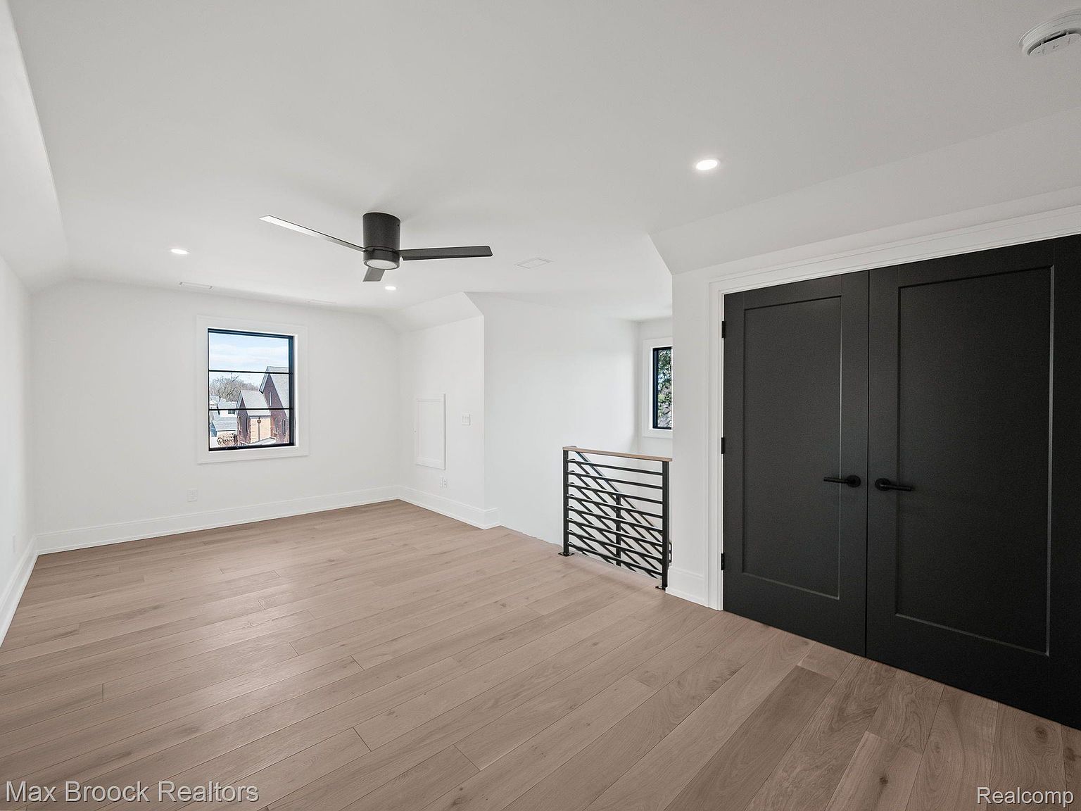 This is an interior shot of a modern bedroom featuring light wood flooring and white walls. A black ceiling fan is mounted on the ceiling, and a window provides natural light. Black double doors are visible on the right, adding a touch of contrast to the room's neutral color palette.