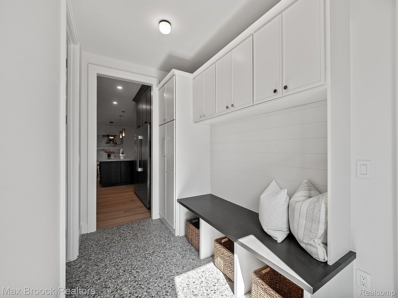 This interior shot showcases a stylish hallway area with built-in storage and seating. White cabinets line the upper wall, complemented by a gray bench with decorative pillows. Woven baskets provide additional storage underneath, and the flooring features a speckled pattern, adding visual interest to the space.