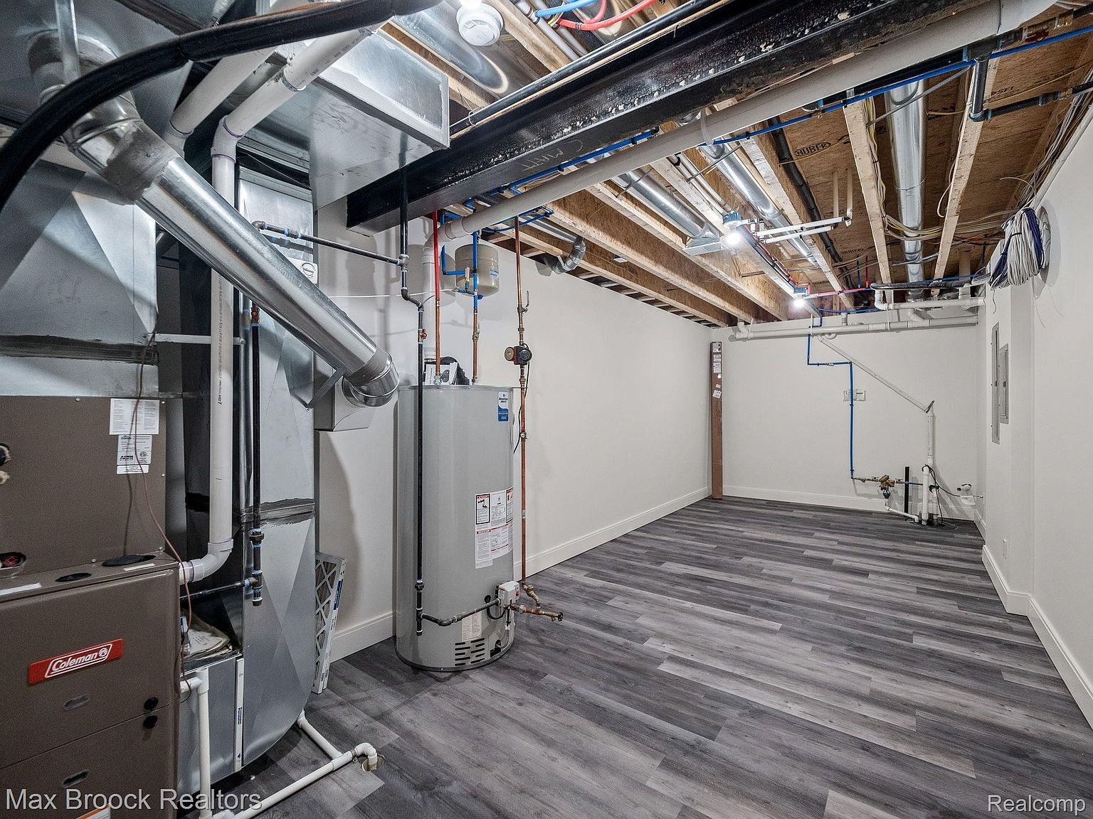 This is a view of a finished basement. The space features gray wood-look flooring, white walls, and an exposed ceiling with visible pipes and ductwork. A water heater and furnace are visible, suggesting a utility area within the finished space.
