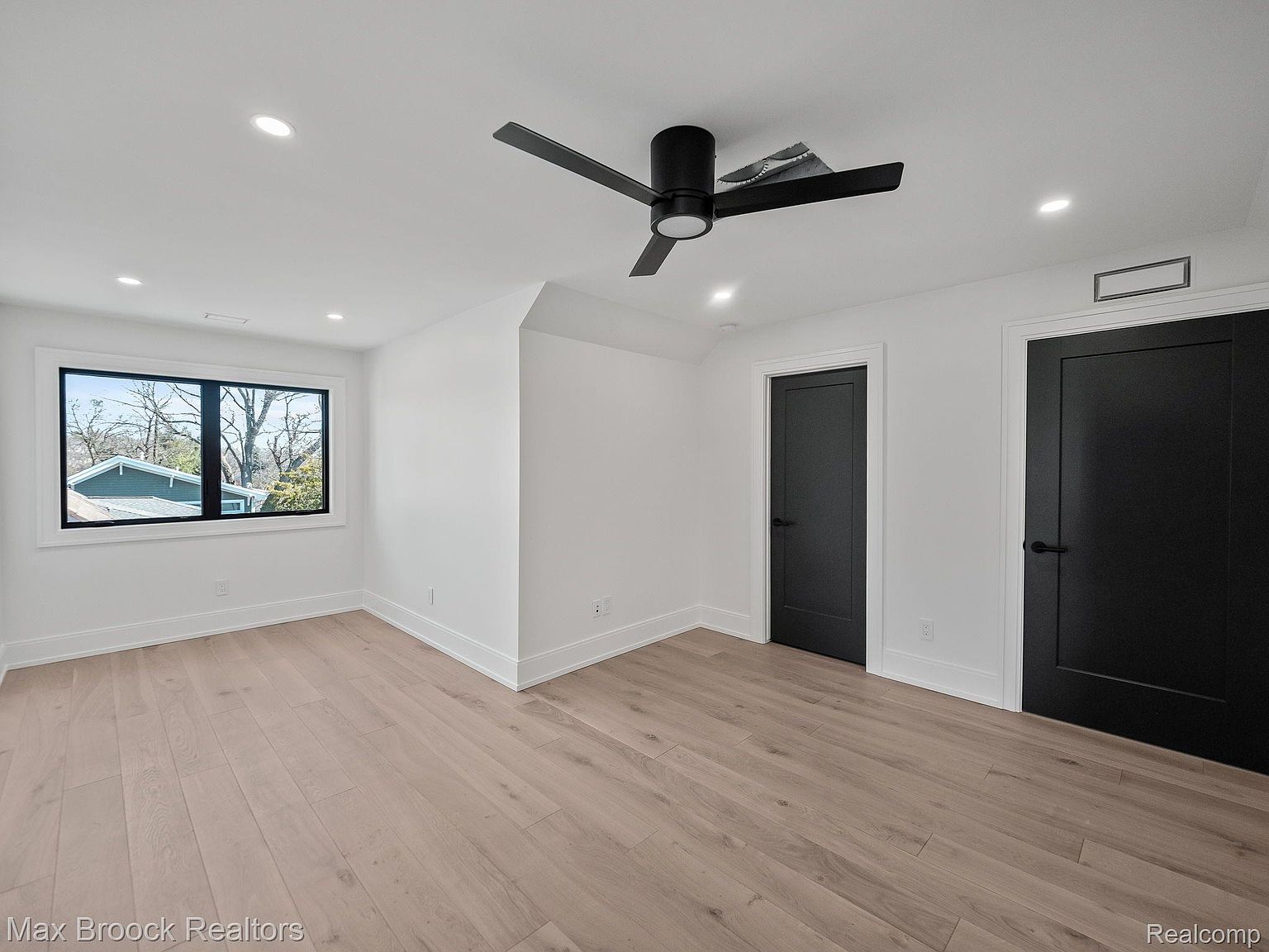 This is an interior shot of a bedroom featuring light hardwood floors and white walls. A black ceiling fan is centered in the room, and a window provides natural light. Two black doors are visible, adding a modern contrast to the space, and the room appears clean and spacious.