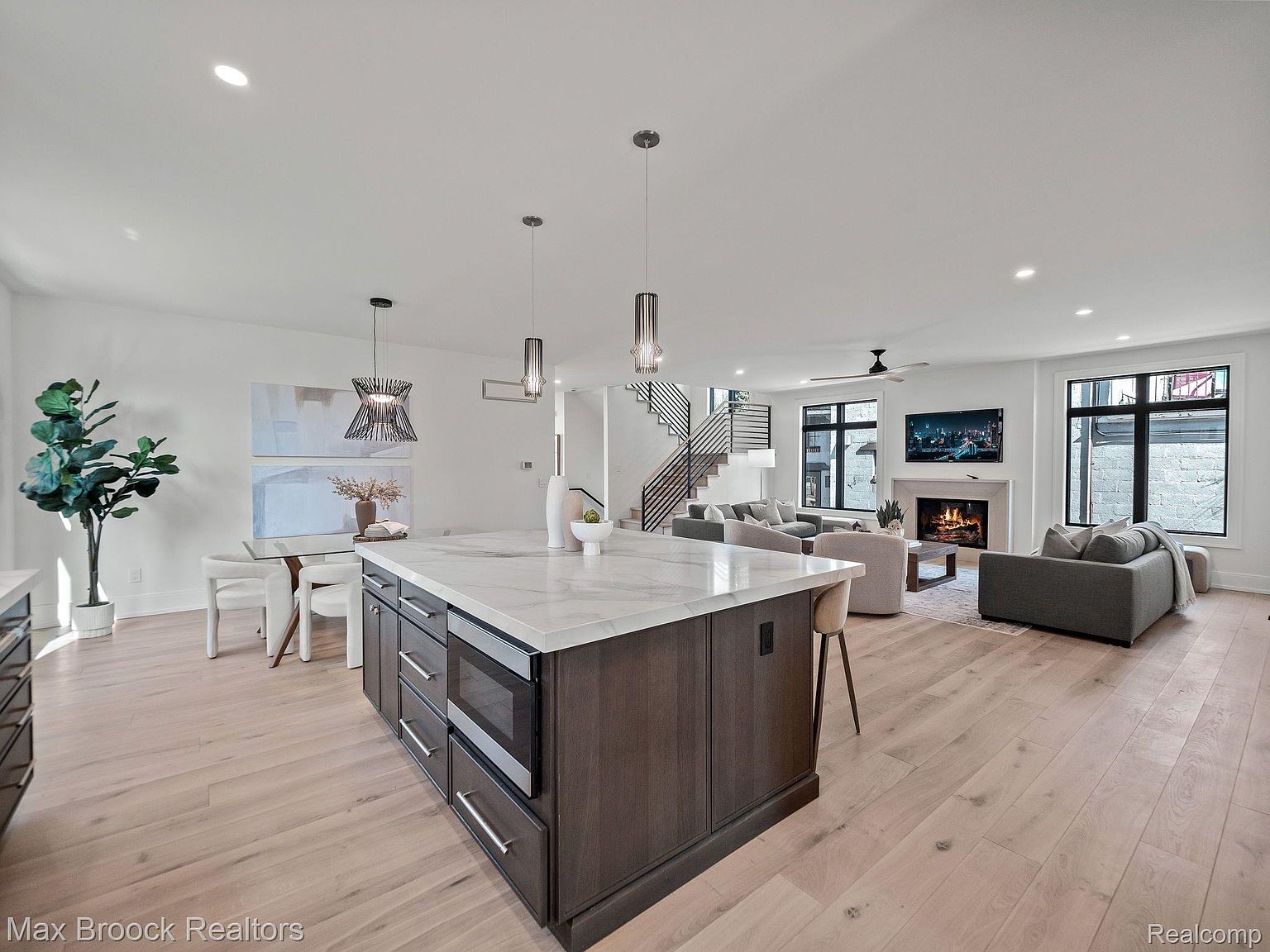 This is a bright, open-concept kitchen and living area. The kitchen features a large island with a marble countertop and dark wood cabinetry, stainless steel appliances, and pendant lighting. The living area includes a fireplace, comfortable seating, and large windows that provide ample natural light, creating a welcoming and modern space.