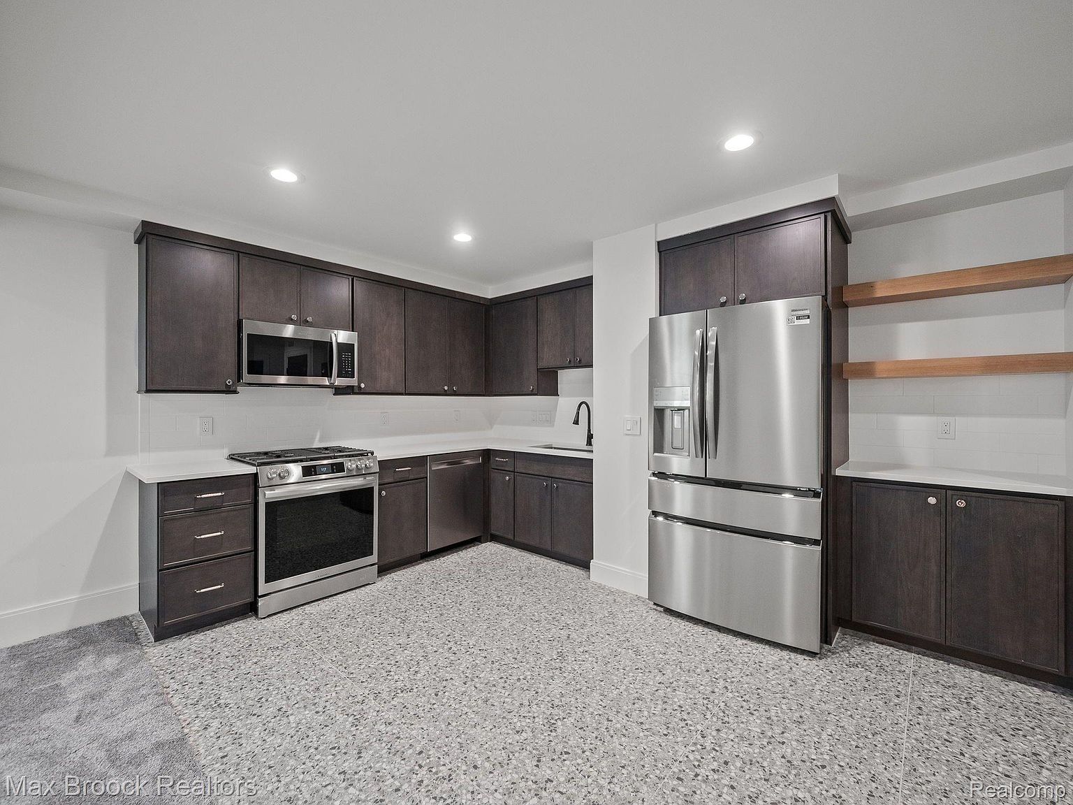 This is a modern kitchen with dark wood cabinetry, stainless steel appliances, and white countertops. The kitchen features a stainless steel refrigerator, a gas range with a stainless steel oven, and a stainless steel microwave. The floor is a speckled gray and white pattern, and the walls are white, creating a clean and contemporary look.