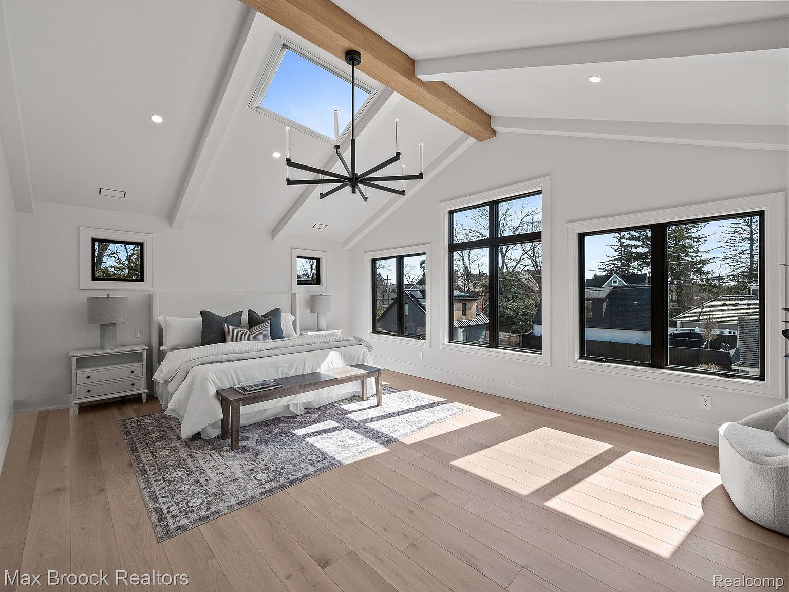 This is a bright and spacious primary bedroom featuring a vaulted ceiling with a skylight and exposed wooden beam. Large windows with black frames offer ample natural light and views of the outdoors. The room is decorated in a neutral color palette with a bed, side tables, and a rug, creating a serene and inviting atmosphere.