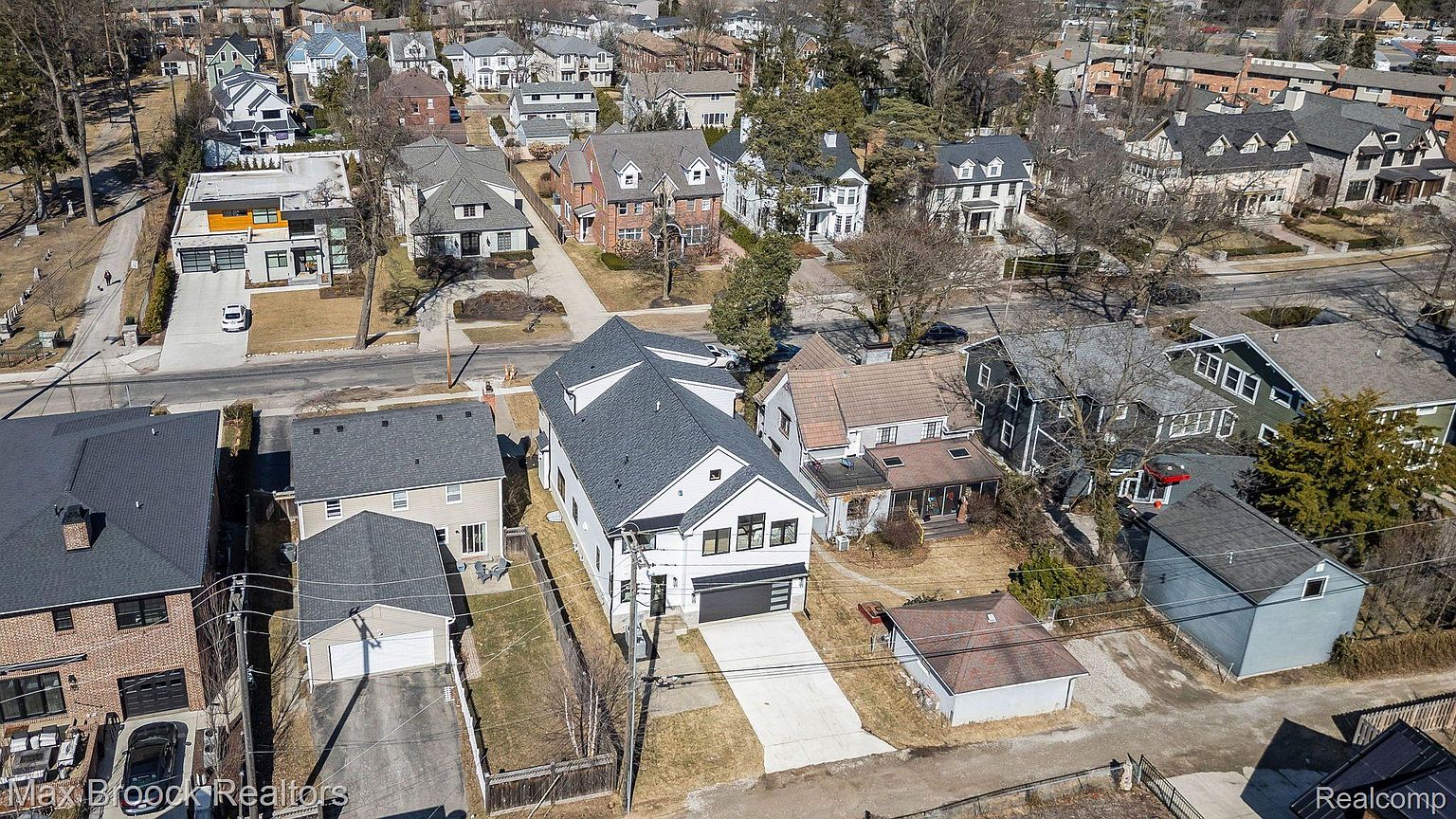 This aerial shot showcases a neighborhood with a mix of architectural styles. The focal point is a modern white house with a dark gray roof and a long driveway. The surrounding homes vary in design, from traditional brick houses to more contemporary structures, all nestled among mature trees and well-maintained lawns.