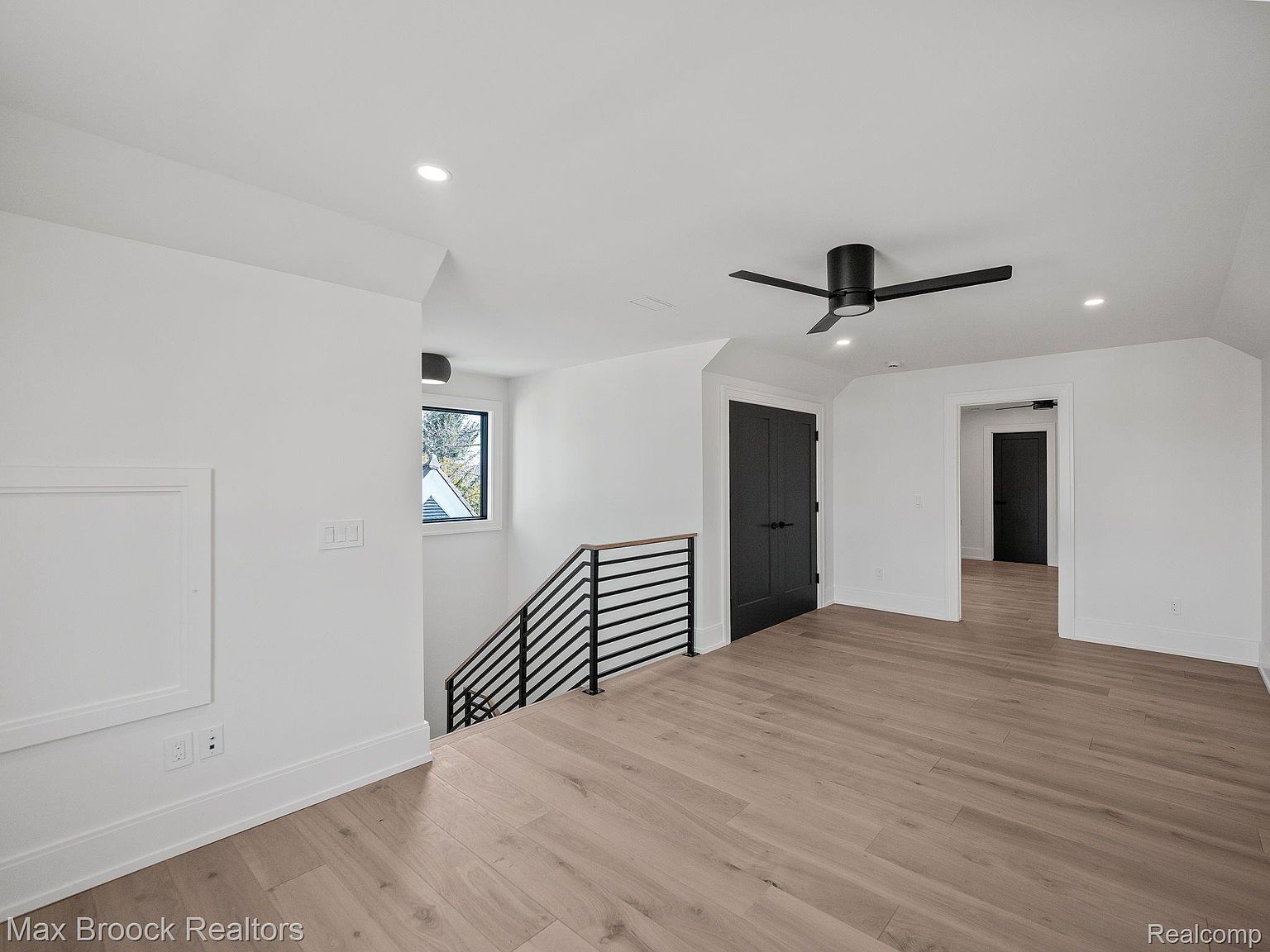 This interior shot showcases a modern hallway and staircase. The hallway features light wood flooring, white walls, and black doors, complemented by a black ceiling fan. A staircase with black railings leads to another level, creating a bright and airy transition space.
