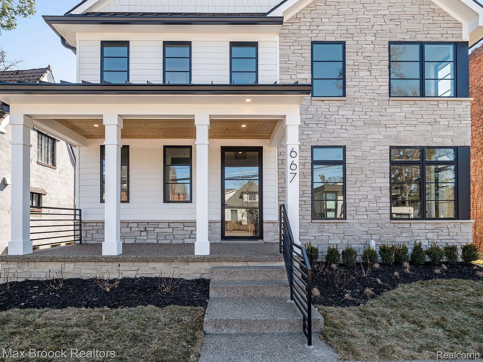 This is a front exterior view of a two-story house featuring a combination of stone and white siding. The house has black-framed windows and a covered front porch with white columns and a wood ceiling. The landscaping includes dark mulch beds and a concrete walkway leading to the front door, creating a welcoming and modern curb appeal.