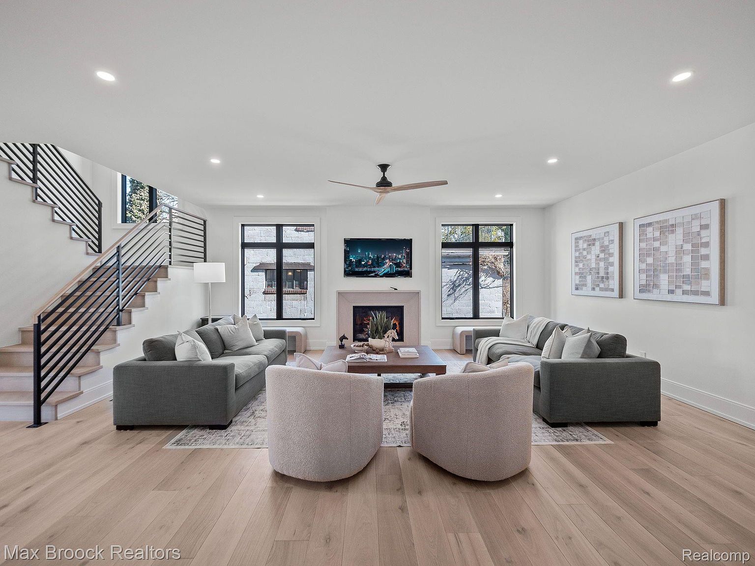 This is a bright and spacious living room featuring hardwood floors, neutral-colored sofas, and two armchairs. A modern fireplace with a television above it serves as a focal point, flanked by large windows. The room is well-lit with recessed lighting and natural light, creating a welcoming and comfortable atmosphere.