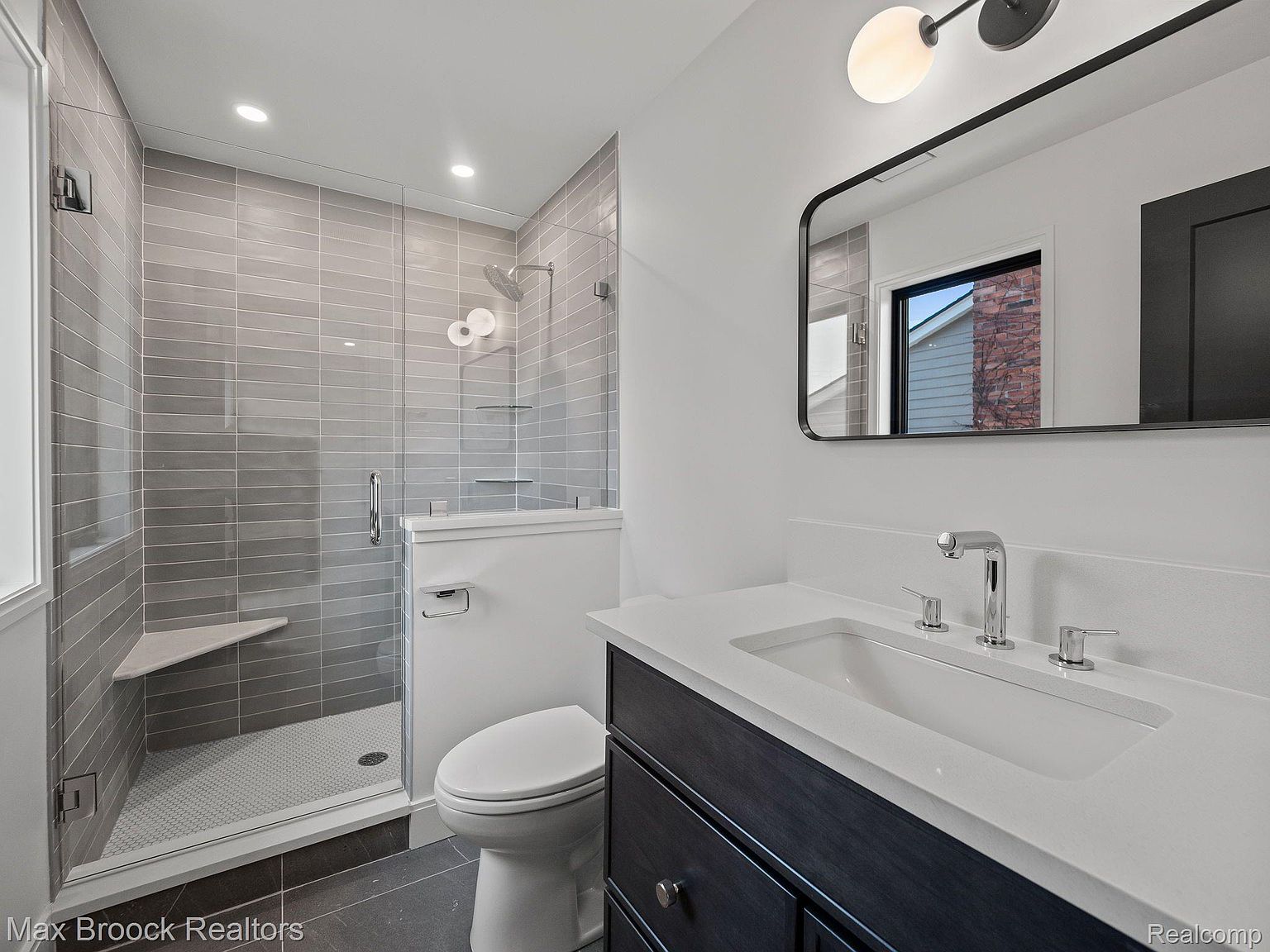 This is a well-lit bathroom featuring a modern design with gray horizontal tiles in the shower area, complemented by a glass door and a built-in bench. The vanity has a dark wood cabinet with a white countertop and a rectangular sink, paired with a sleek mirror and modern lighting fixture. The toilet is white and positioned next to the shower, creating a functional and stylish space.