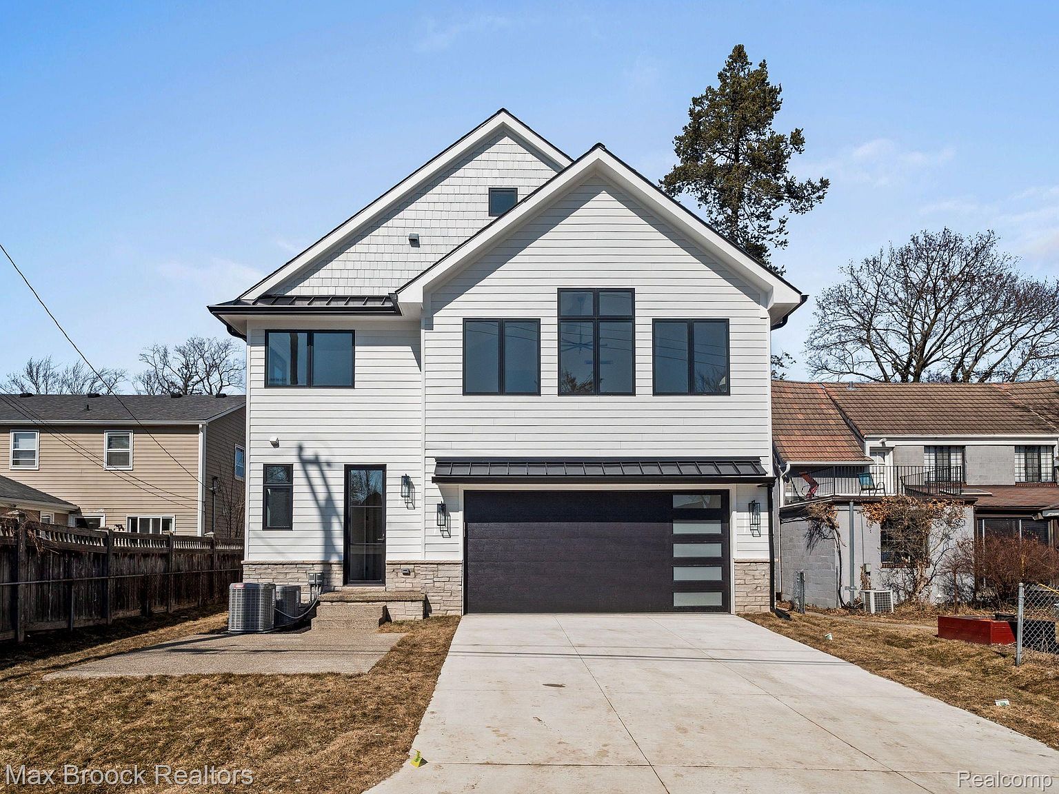 This is a front view of a modern two-story house with white siding and black-framed windows. The house features a dark gray garage door with modern window accents and a concrete driveway. The landscaping is minimal, with a focus on the architectural design of the home.