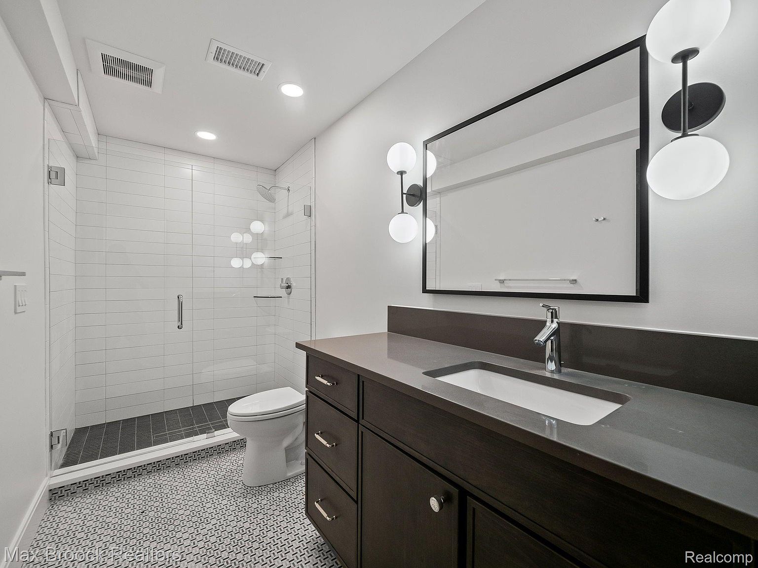 This is a well-lit bathroom featuring a modern design with a dark wood vanity, gray countertop, and a rectangular sink. A large mirror with unique sconces hangs above the vanity. The bathroom also includes a glass-enclosed shower with white subway tile and a toilet, all set against a patterned tile floor.