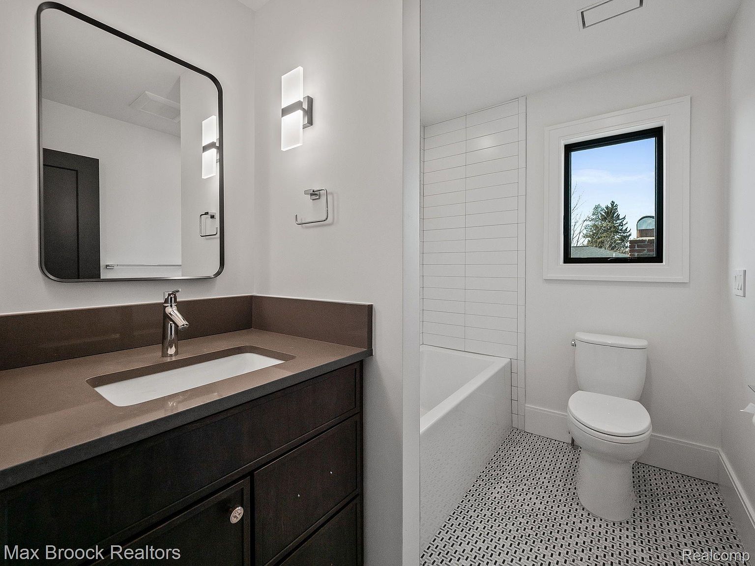 This is a well-lit bathroom featuring a dark wood vanity with a light countertop and a rectangular sink. A modern mirror hangs above the vanity, and contemporary light fixtures adorn the wall. The bathroom also includes a toilet and a bathtub with white subway tile, complemented by a patterned black and white tile floor, creating a clean and stylish space.