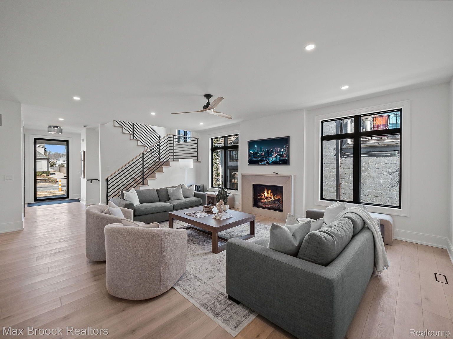This is a bright and airy living room featuring a modern design with hardwood floors, a neutral color palette, and ample natural light. The room is furnished with comfortable seating arrangements, including sofas and armchairs, centered around a coffee table and a fireplace with a mounted television above. A staircase with modern railings is visible in the background, adding to the contemporary aesthetic.