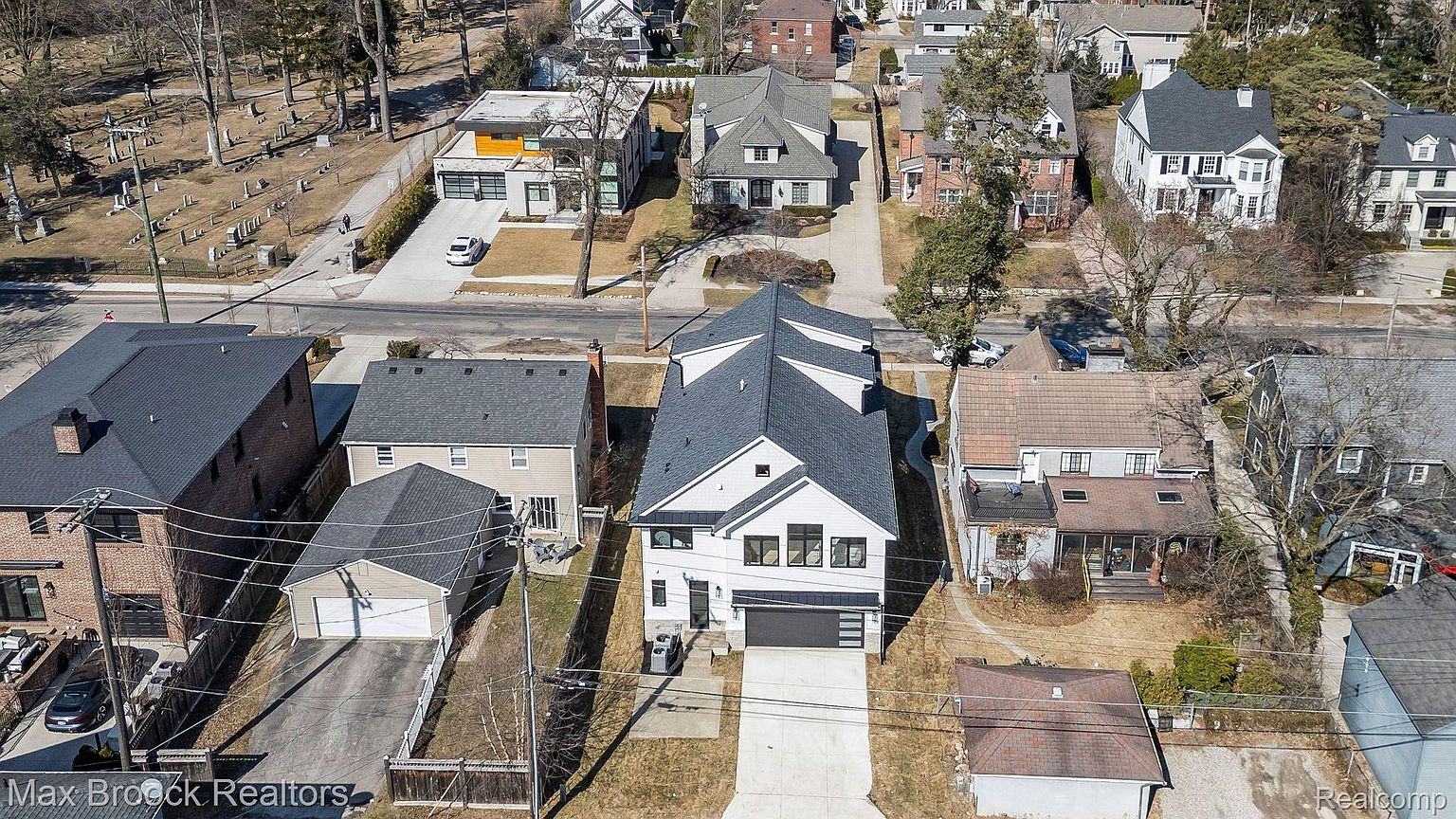 This aerial shot showcases a neighborhood with a mix of architectural styles, including modern and traditional homes. The focal point is a newly constructed house with a dark gray roof, white exterior, and a modern garage door. The surrounding properties feature well-maintained yards and mature trees, creating a desirable residential setting.