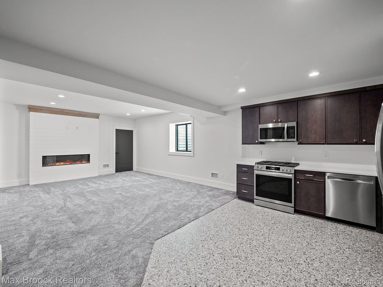 This is an interior shot of a finished basement, featuring a living area with a modern fireplace and a kitchenette. The living area has gray carpeting, while the kitchenette has a speckled tile floor. The kitchenette includes dark wood cabinets, stainless steel appliances, and a white countertop, creating a functional and stylish space.