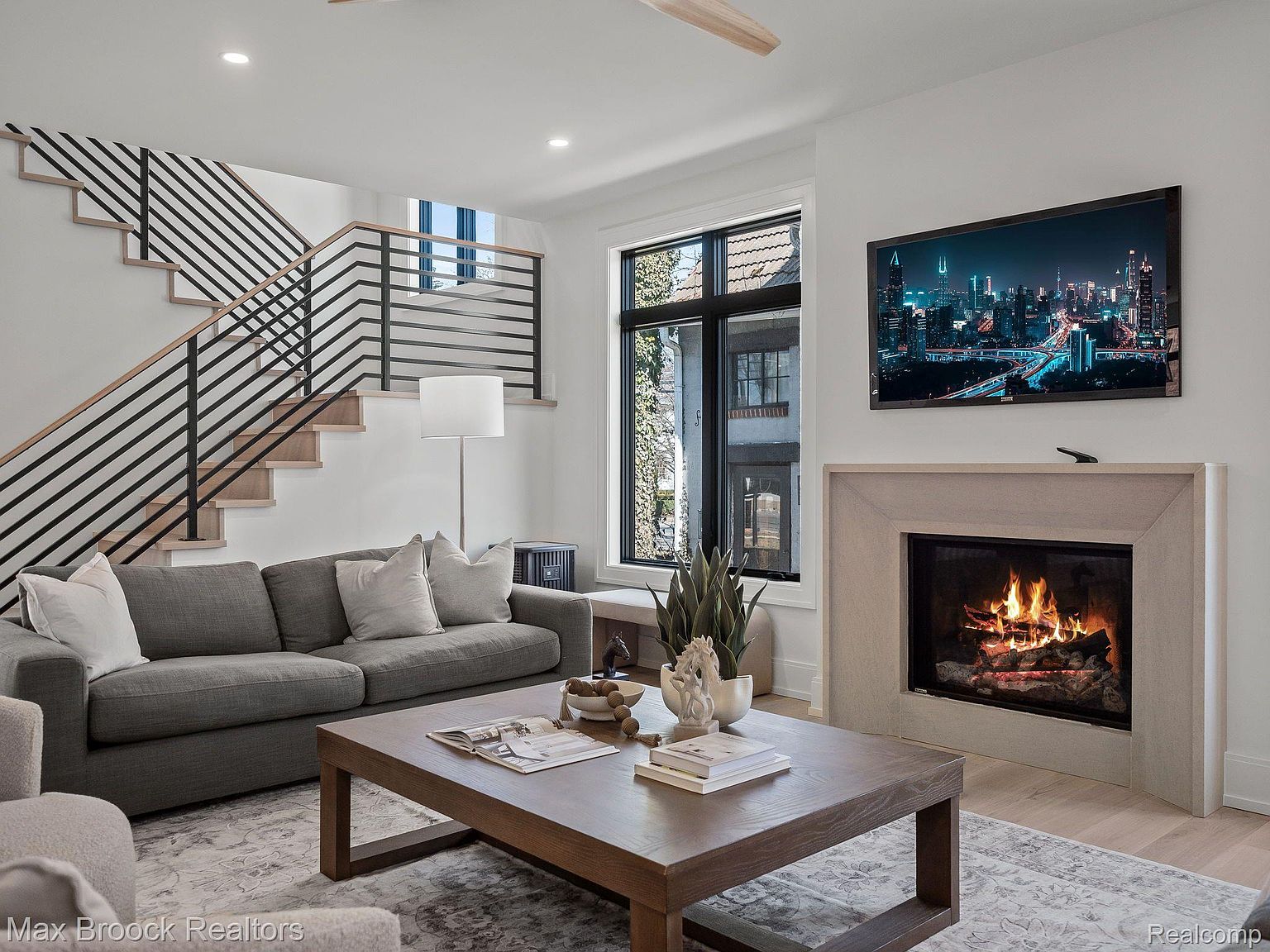 This is an interior shot of a modern living room featuring a gray sofa with throw pillows, a large wooden coffee table, and a fireplace with a mounted television above it. A staircase with black railings is visible on the left, and a large window provides natural light. The room is decorated in neutral tones, creating a cozy and inviting atmosphere.