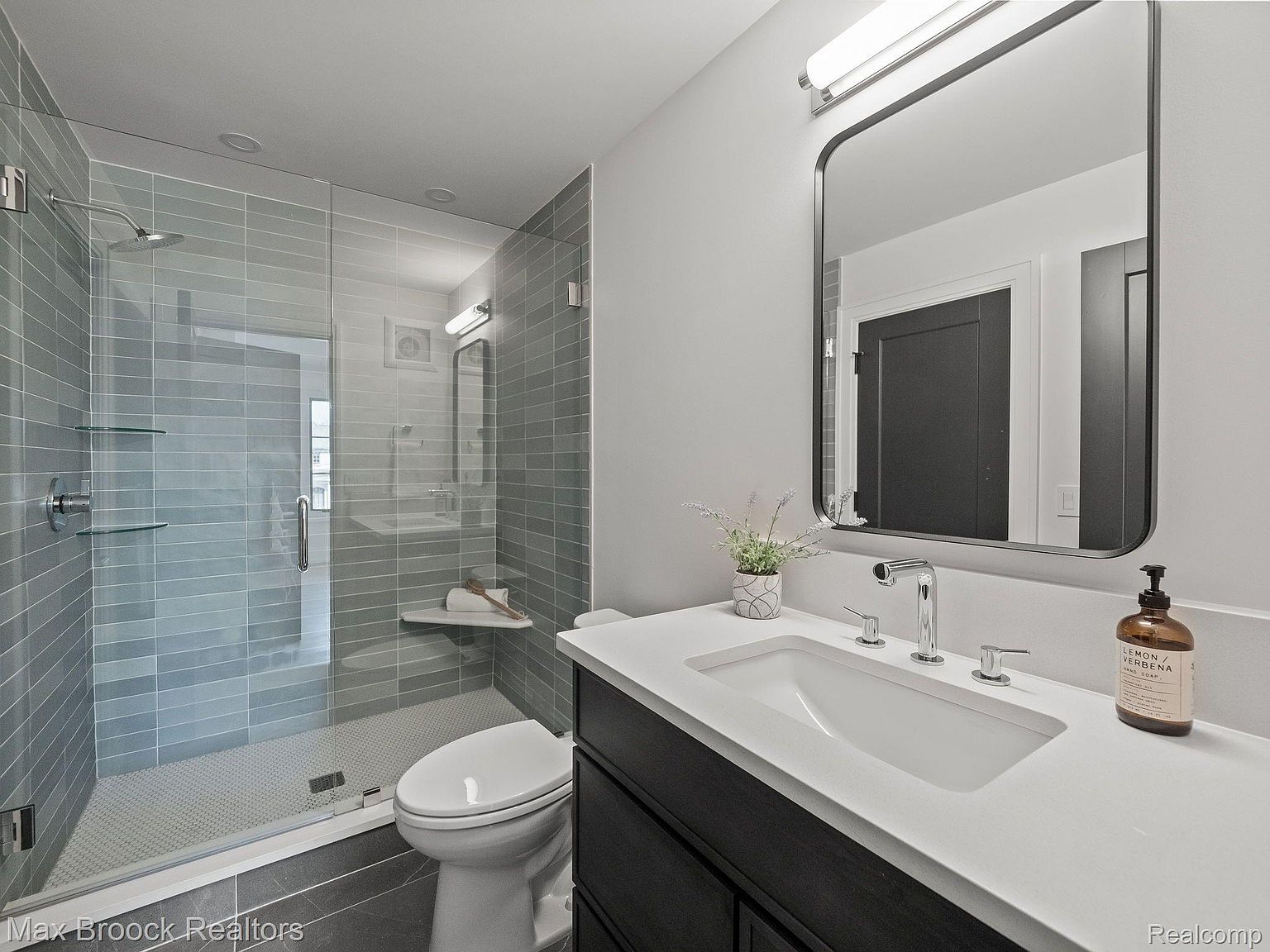 This is a modern bathroom featuring a glass-enclosed shower with gray tiling and a built-in bench. The vanity has a white countertop, a dark wood cabinet, and a rectangular mirror above. The overall design is clean and contemporary, creating a spa-like atmosphere.