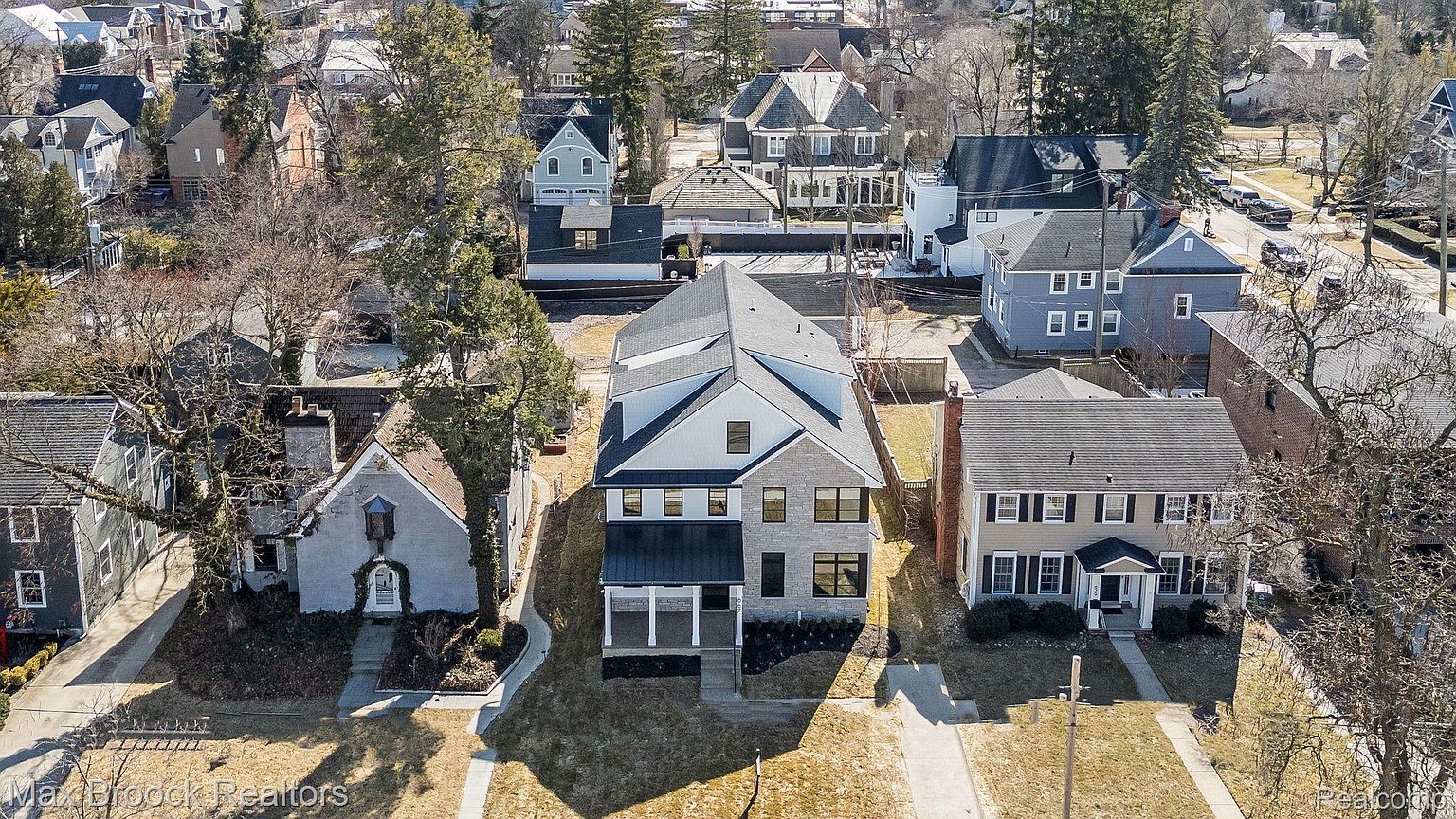 This aerial view showcases a well-maintained residential property with a modern architectural style. The house features a combination of stone and white siding, a dark metal roof, and a welcoming front porch. The surrounding neighborhood is characterized by mature trees and a mix of traditional and contemporary homes, creating a desirable and established community.