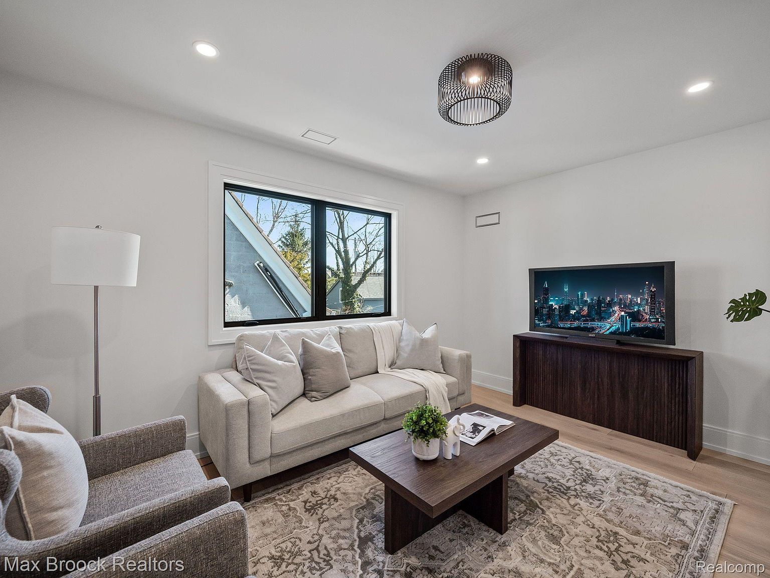 This is a bright and modern living room featuring a neutral color palette. The room is furnished with a light-colored sofa, a gray armchair, a dark wood coffee table, and a console table supporting a large television. A large window provides natural light, and the room is decorated with a patterned rug and contemporary lighting fixtures.