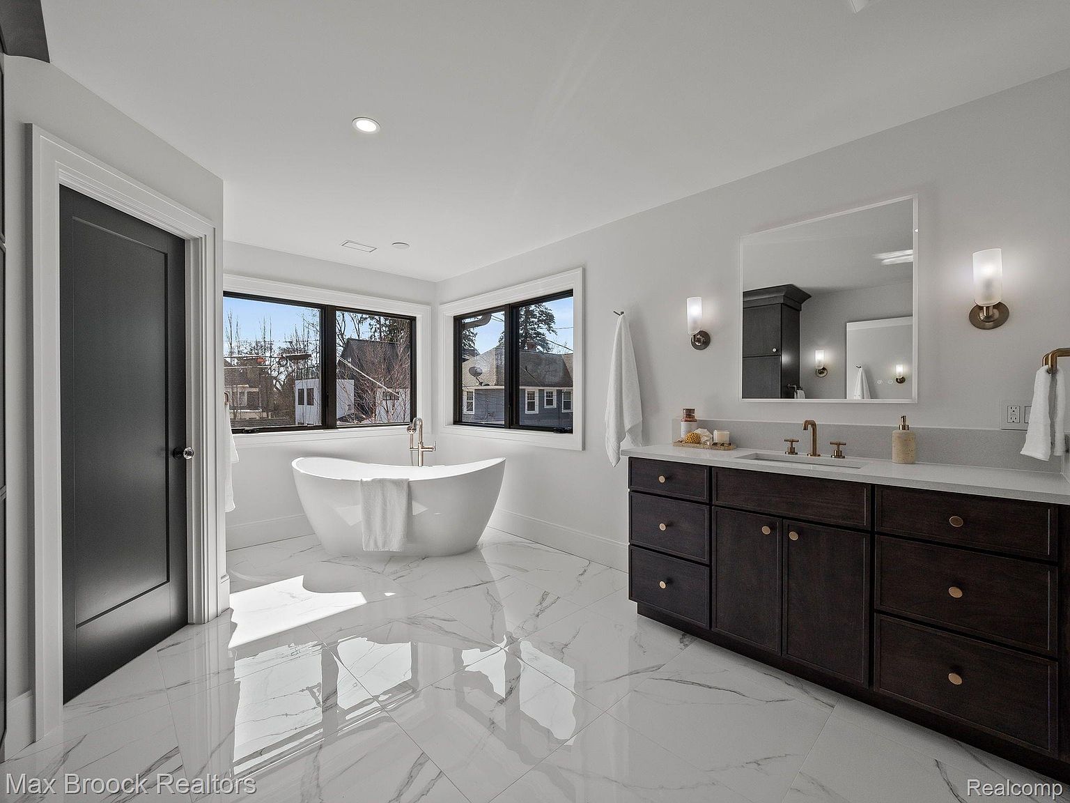 This is a luxurious primary bathroom featuring a freestanding white bathtub positioned in front of a large window, offering natural light. A dark wood vanity with a white countertop and gold fixtures adds a touch of elegance. The flooring is a polished white marble, enhancing the room's bright and spacious feel.