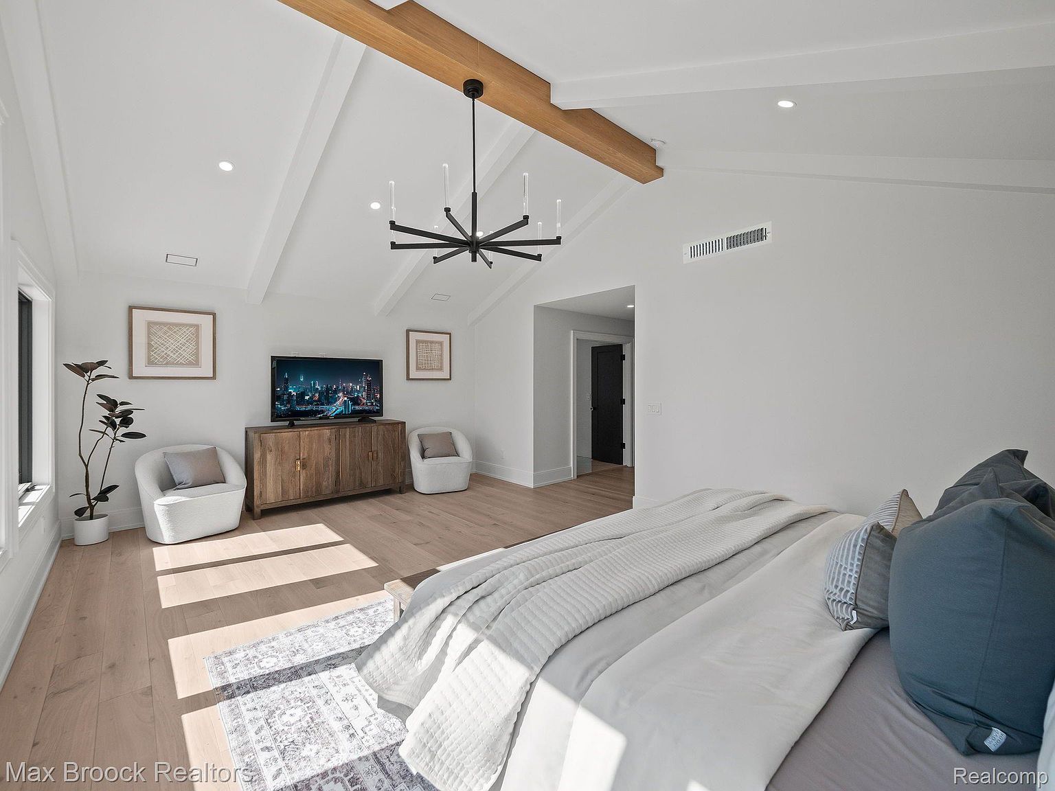 This is a bright and airy primary bedroom featuring a vaulted ceiling with exposed wooden beams and a modern chandelier. The room is furnished with a wooden media console, two armchairs, and a large bed with plush bedding. Natural light floods the space, highlighting the light wood flooring and neutral color palette, creating a serene and inviting atmosphere.
