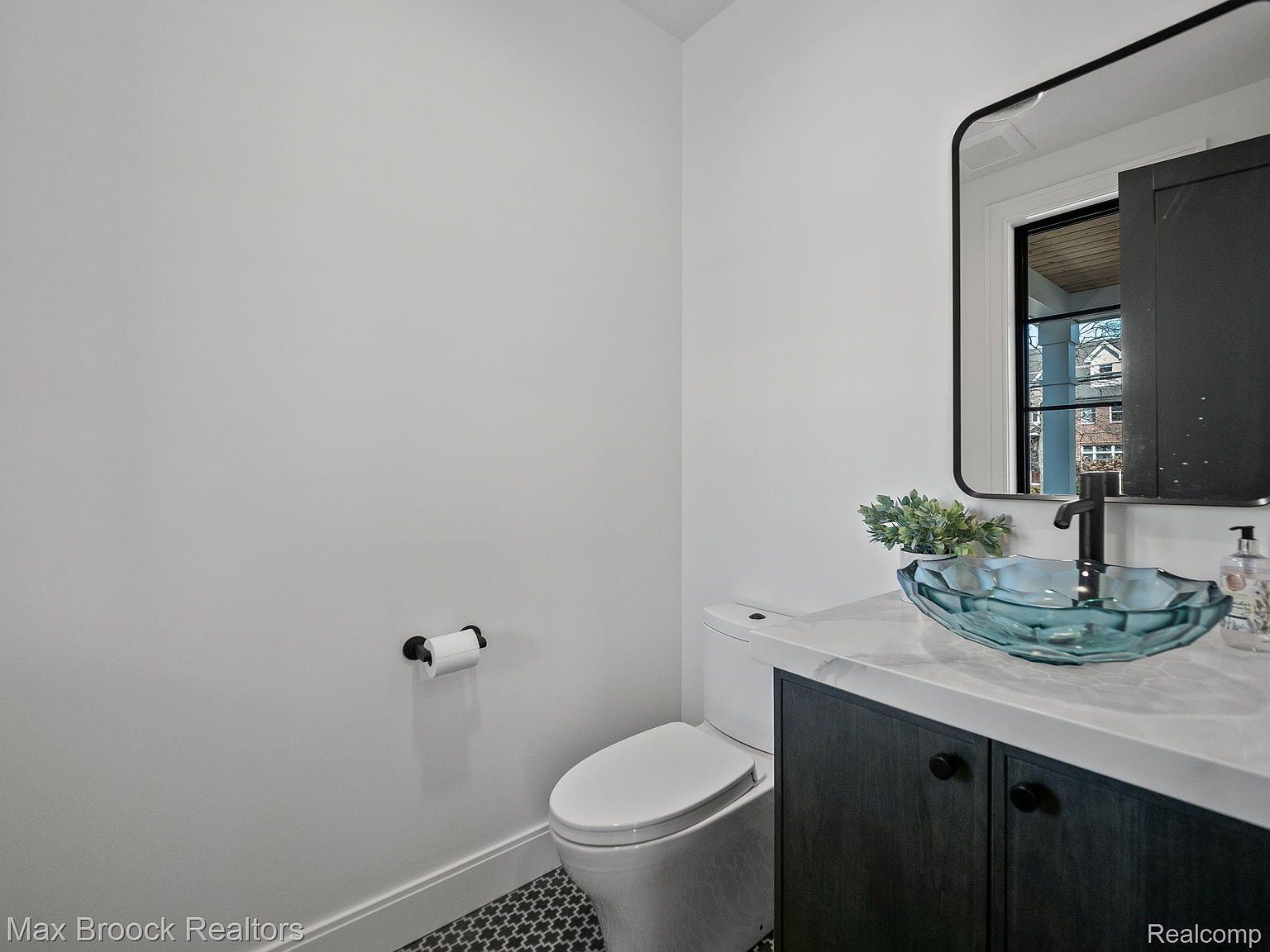 This is a well-lit guest bathroom featuring a modern design. The room includes a toilet, a vanity with a unique blue glass vessel sink, a black faucet, and a black-framed mirror. The floor is tiled with a geometric pattern, and the walls are painted a neutral color, creating a clean and stylish space.