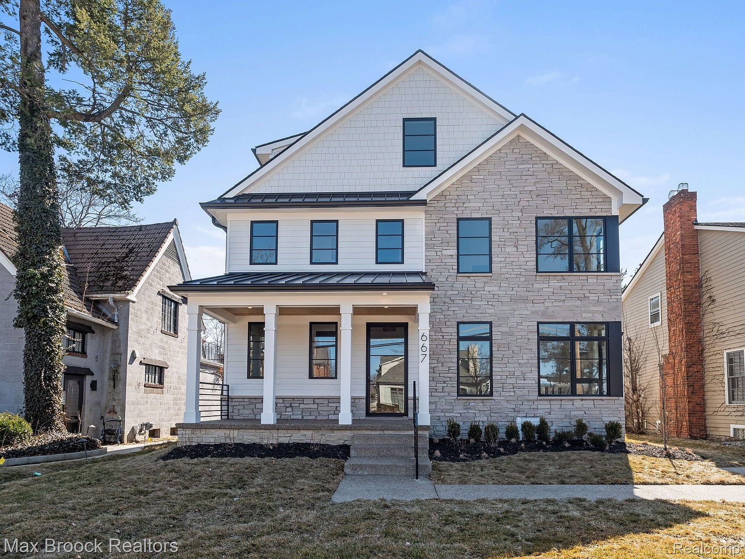 This is a front view of a two-story house with a combination of white siding and stone facade. The house features black-framed windows, a covered porch with white columns, and a well-maintained lawn. The architectural style is modern and elegant, creating a welcoming and upscale impression.