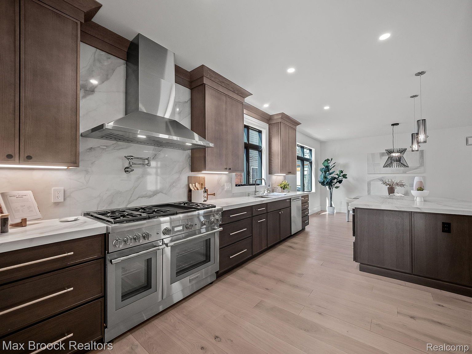 This is a well-lit kitchen featuring dark wood cabinetry, stainless steel appliances, and light-colored hardwood floors. A large stainless steel range hood is positioned above the stove, and a kitchen island is visible in the background. The perspective is a wide shot, capturing the layout and design elements of the kitchen.