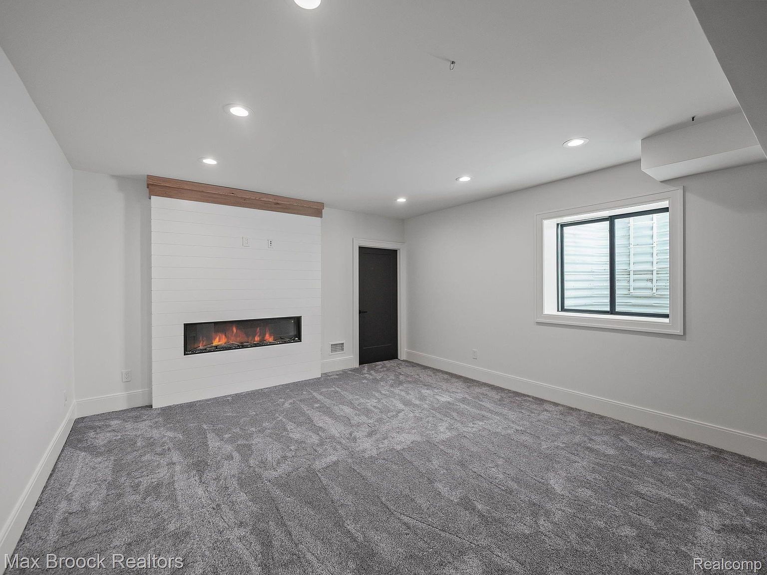 This is an interior shot of a living room featuring a modern fireplace with a wooden mantel and a white shiplap surround. The room has gray carpeting and a window providing natural light. The overall impression is clean, contemporary, and inviting.