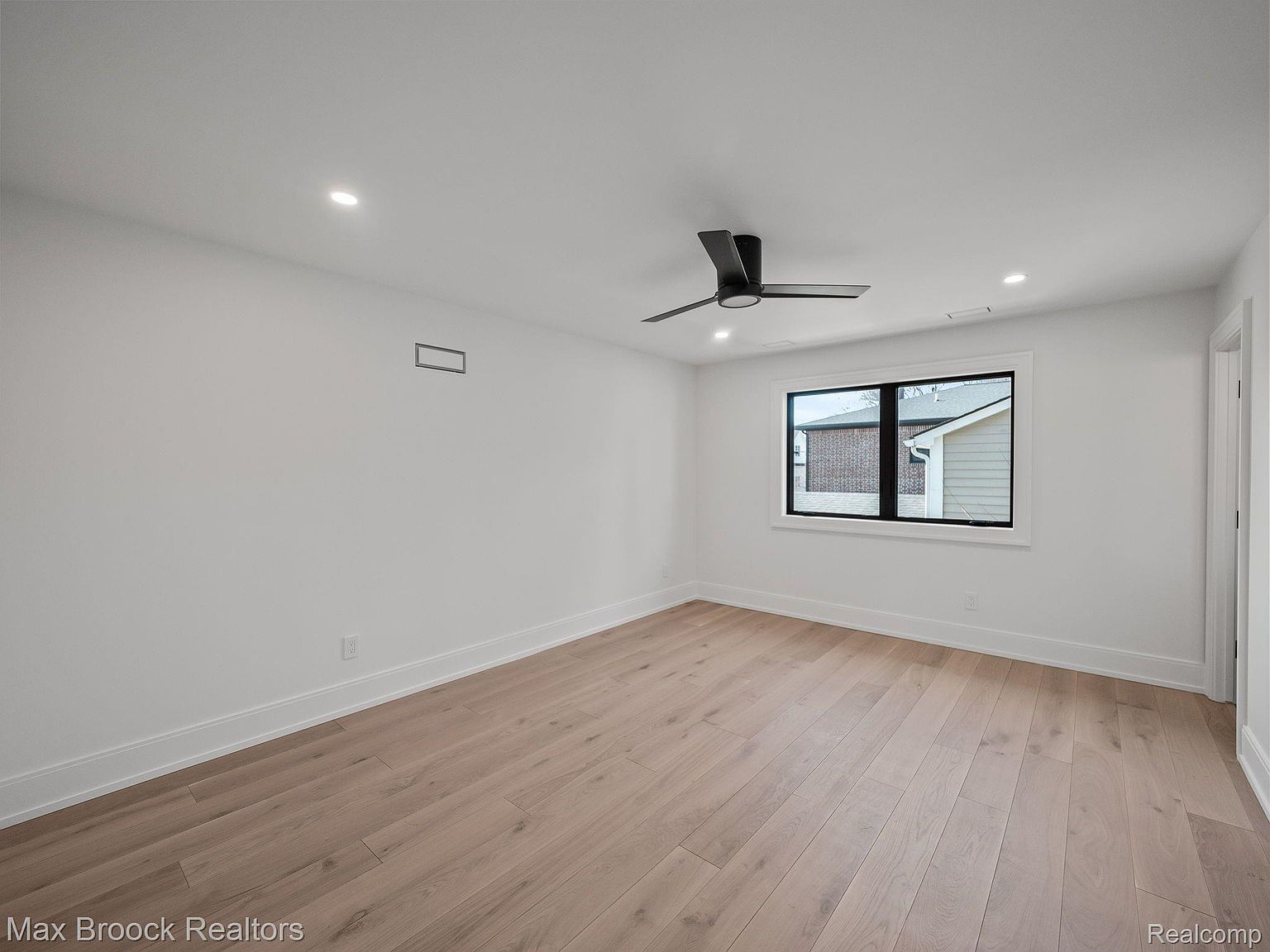 This is a bright and airy bedroom featuring light hardwood floors and white walls. A modern ceiling fan and a large window provide ample light and ventilation. The room is clean and minimalist, offering a blank canvas for potential buyers to envision their own style.
