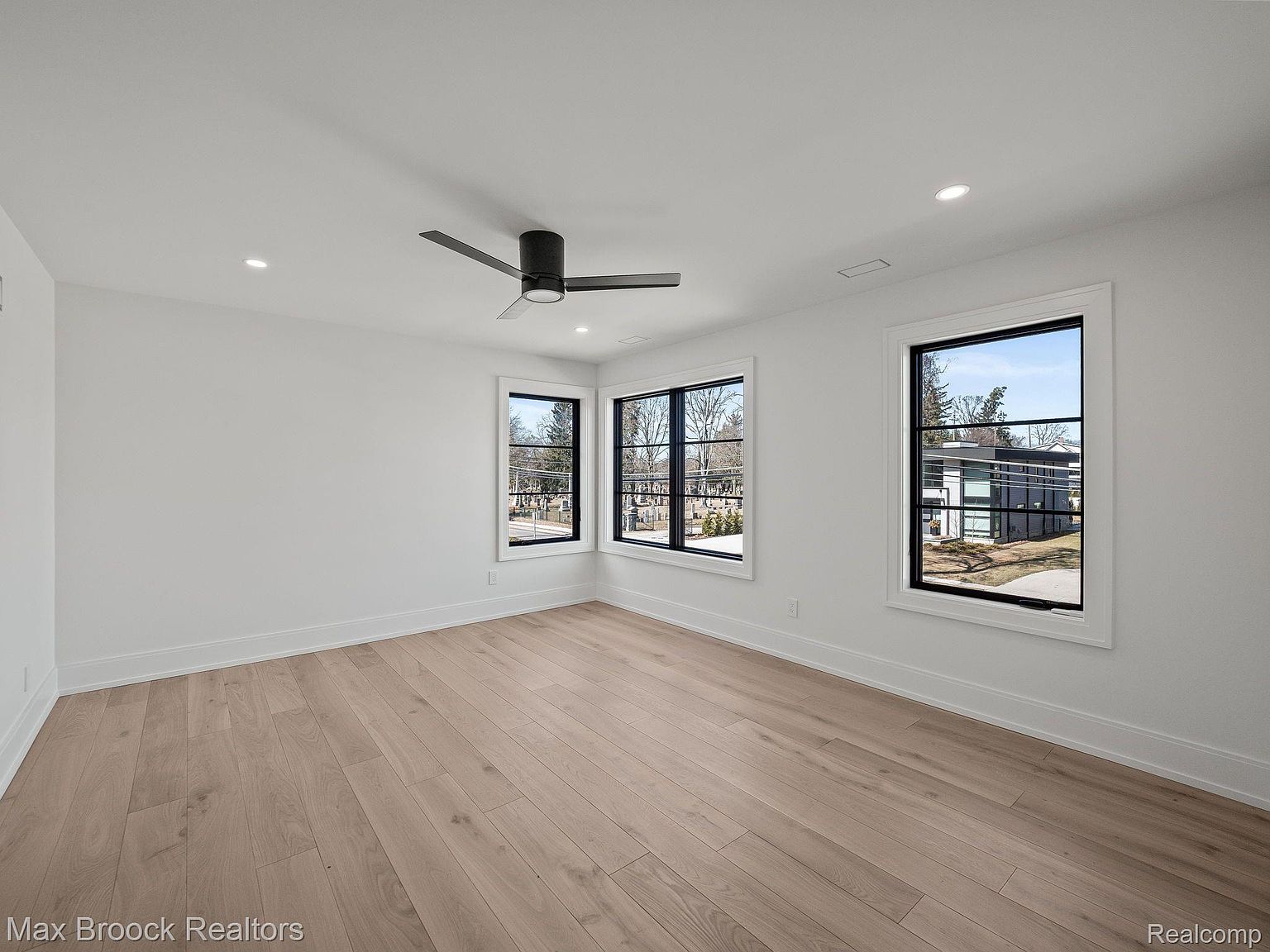 This is a bright and spacious primary bedroom featuring light hardwood flooring, white walls, and modern black-framed windows that provide ample natural light. A sleek black ceiling fan adds a contemporary touch. The room is clean and minimalist, offering a blank canvas for potential buyers to envision their personal style.
