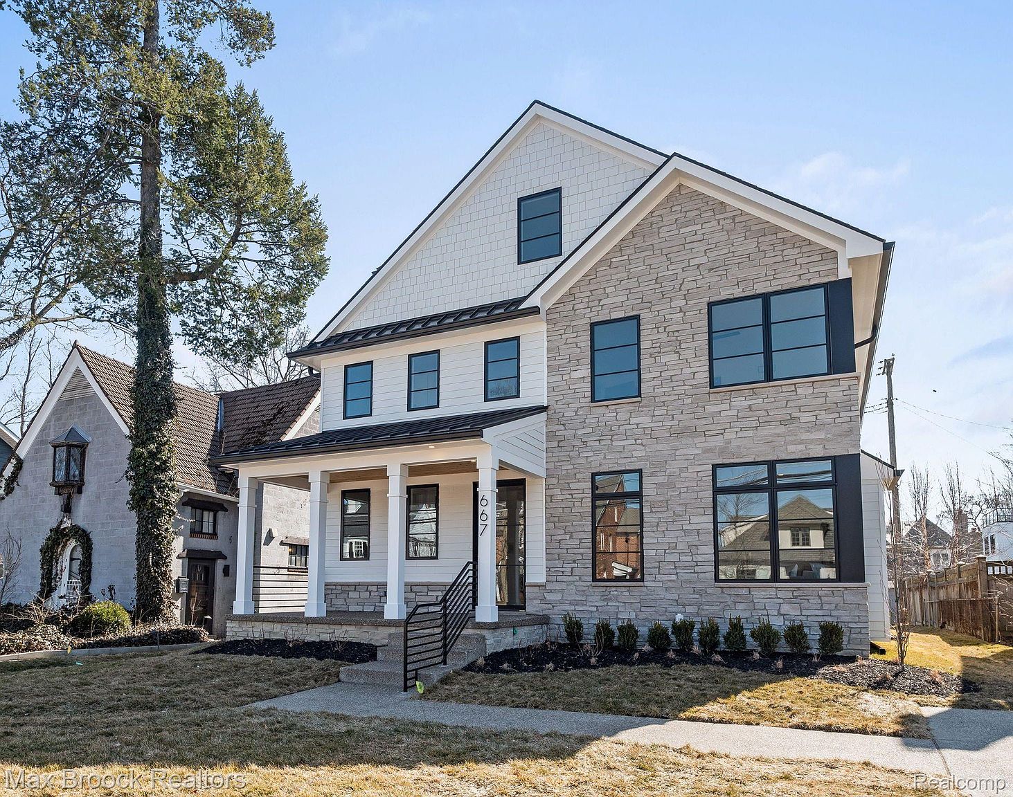 This is a front exterior view of a two-story house featuring a combination of white siding and stone facade. The house has black-framed windows and a covered front porch with white columns. The landscaping includes neatly trimmed bushes and a well-maintained lawn, contributing to the property's curb appeal.