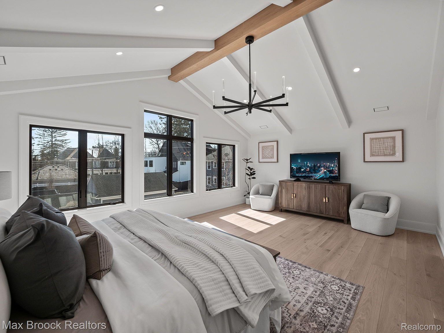 This is a bright and airy primary bedroom featuring a vaulted ceiling with exposed beams and a modern chandelier. Three large windows provide ample natural light and views of the neighborhood. The room is furnished with a bed, two armchairs, a wooden cabinet with a TV, and a patterned area rug, creating a comfortable and stylish retreat.