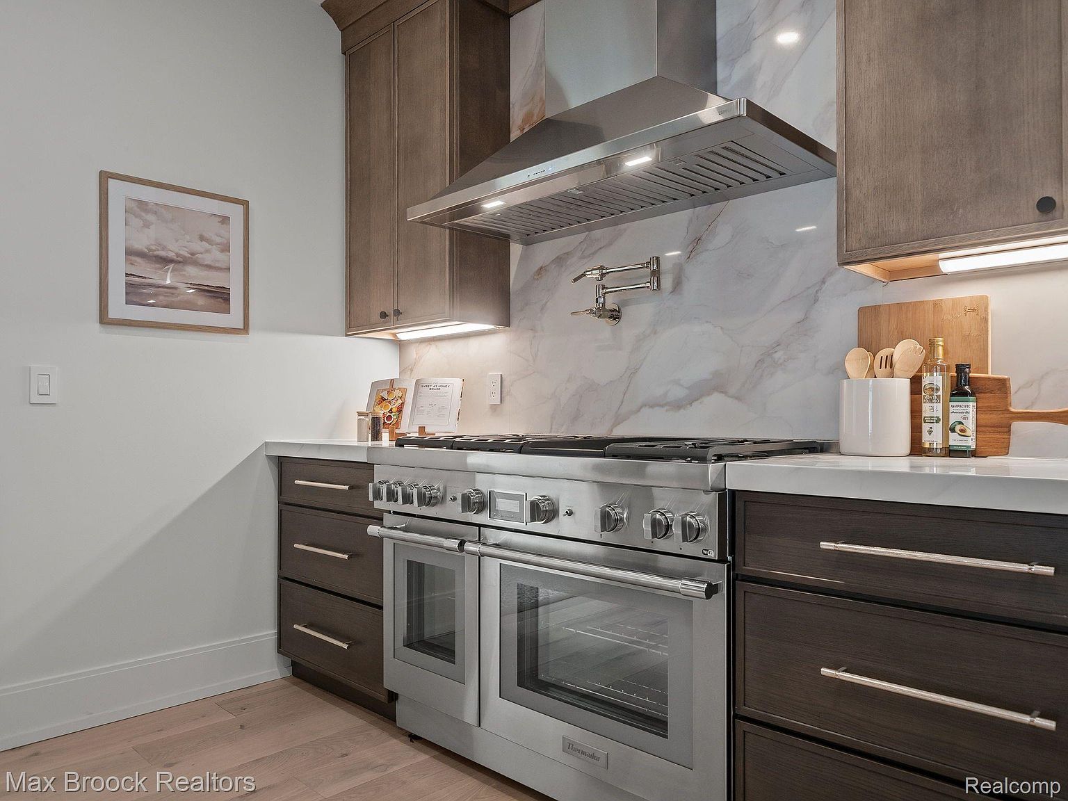 This is a well-appointed kitchen featuring dark wood cabinetry with sleek, modern hardware and stainless steel appliances, including a large range with a double oven and a stainless steel range hood. The countertops are light-colored, possibly marble or quartz, and the backsplash is a matching marble slab. The overall impression is one of luxury and functionality, perfect for a home chef.