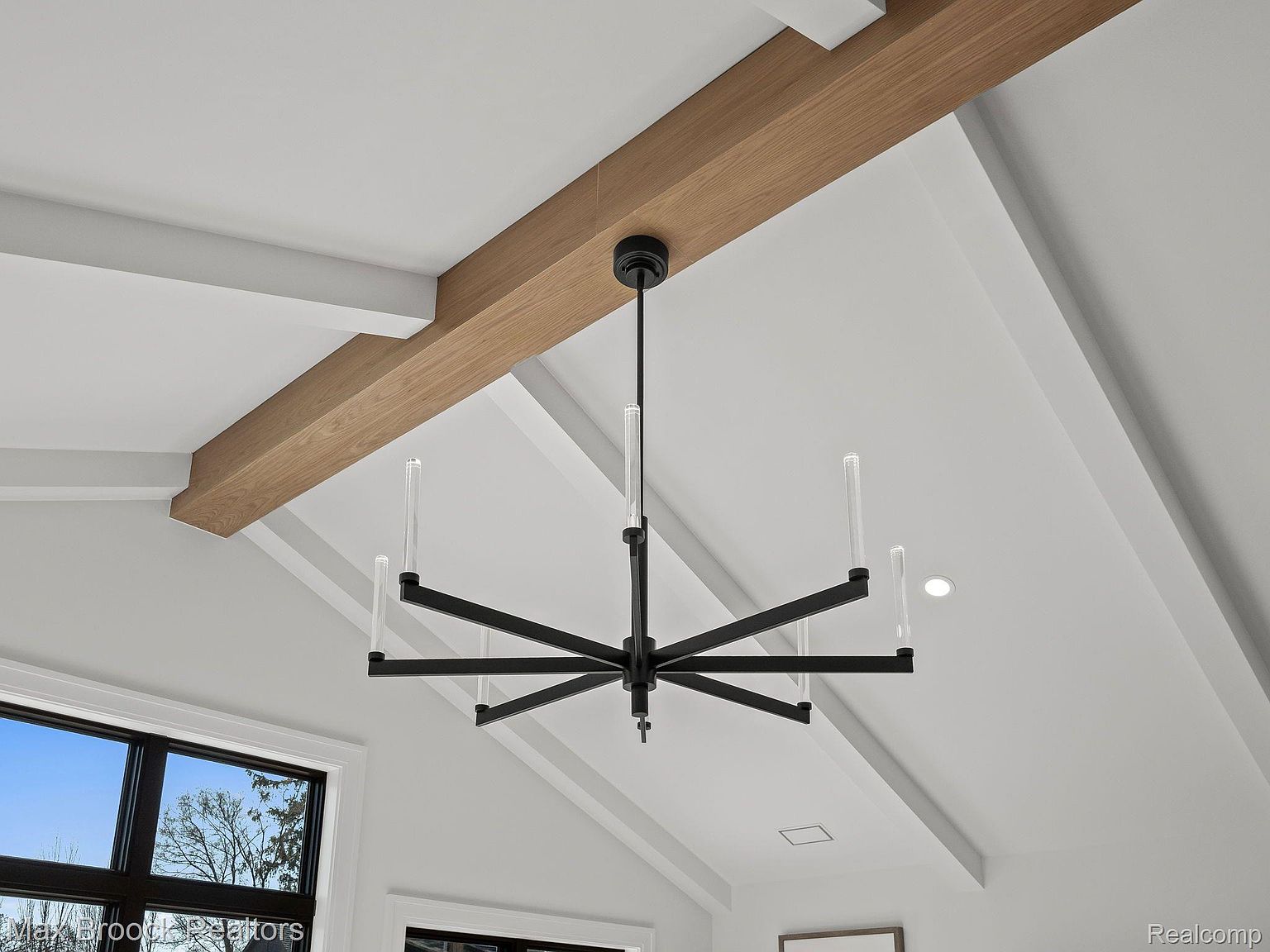 The image showcases an interior space with a modern chandelier hanging from a high, vaulted ceiling. The ceiling features wooden beams, adding a touch of warmth to the otherwise white space. The overall impression is clean, bright, and stylish, suggesting a well-designed and maintained home.