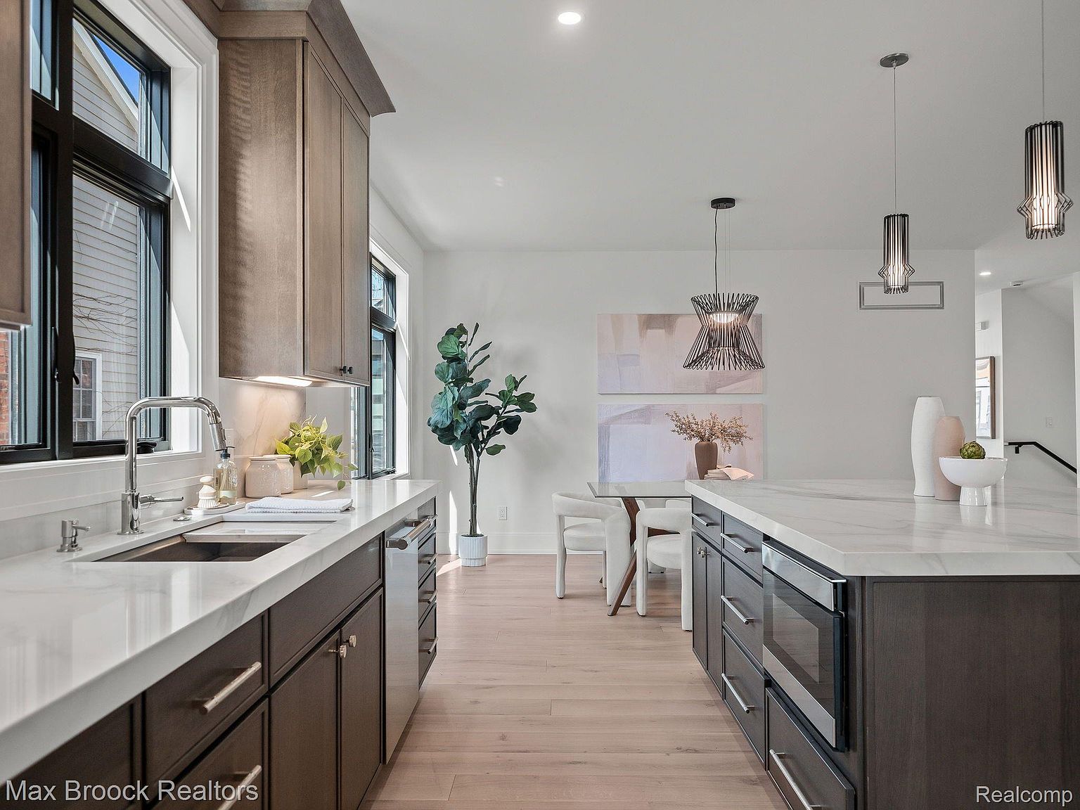 This is a bright and modern kitchen featuring dark wood cabinetry, white marble countertops, and stainless steel appliances. The kitchen island provides ample counter space and includes built-in appliances. The space is well-lit with natural light from the windows and pendant lighting above the island, creating a clean and inviting atmosphere.