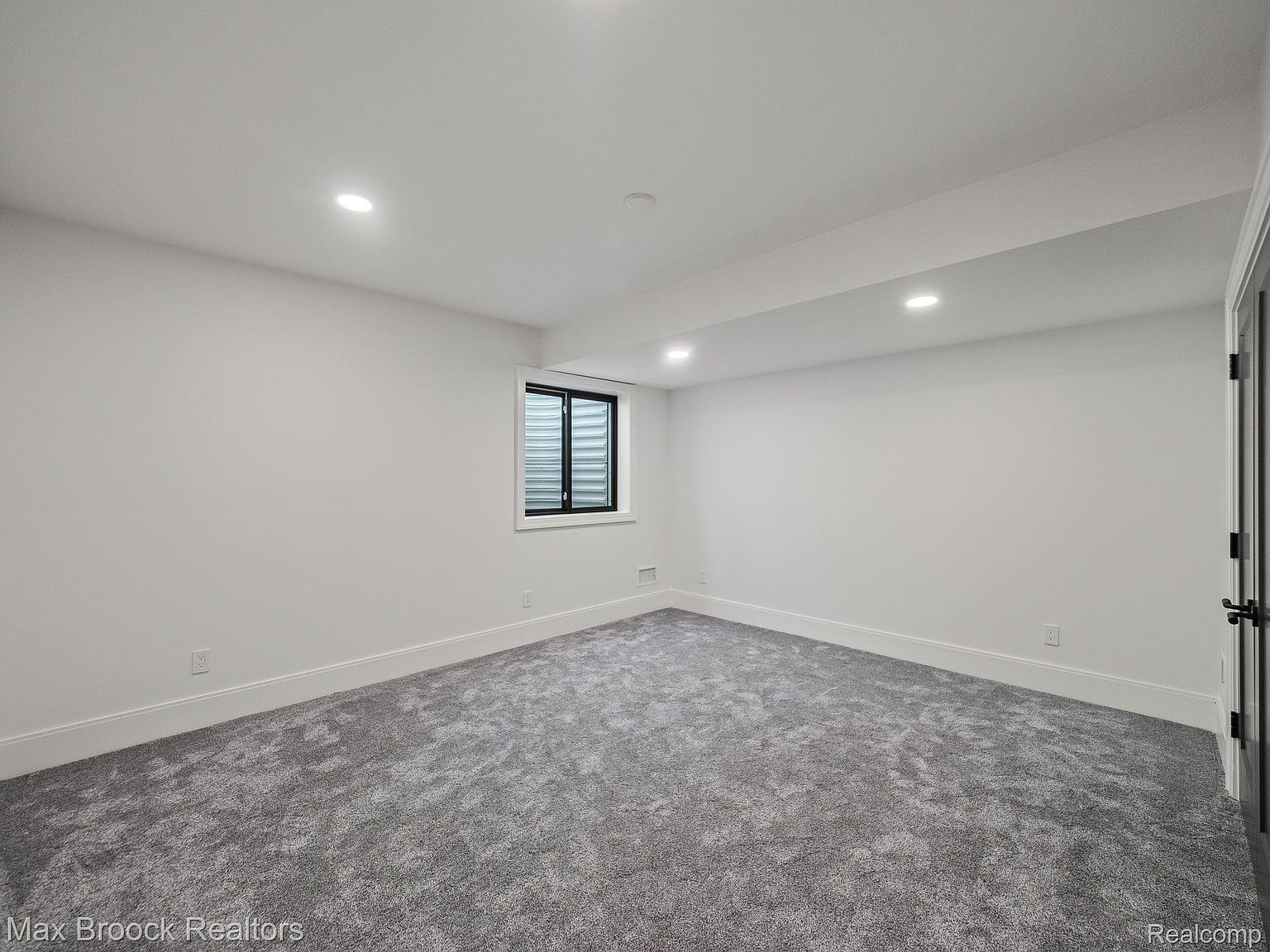 The image showcases a finished basement with gray carpet and white walls. A small window provides natural light, and recessed lighting illuminates the space. The room appears clean and spacious, suitable for various uses such as a recreation area or additional living space.