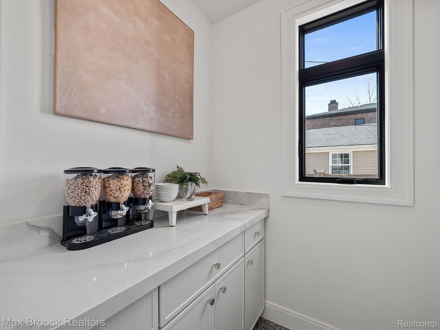 This is an interior shot of a pantry area featuring a white countertop with marble-like veining and white cabinets below. Three cereal dispensers sit on the counter, along with a small plant and bowls on a white stand. A window provides natural light, and a large piece of artwork hangs on the wall.