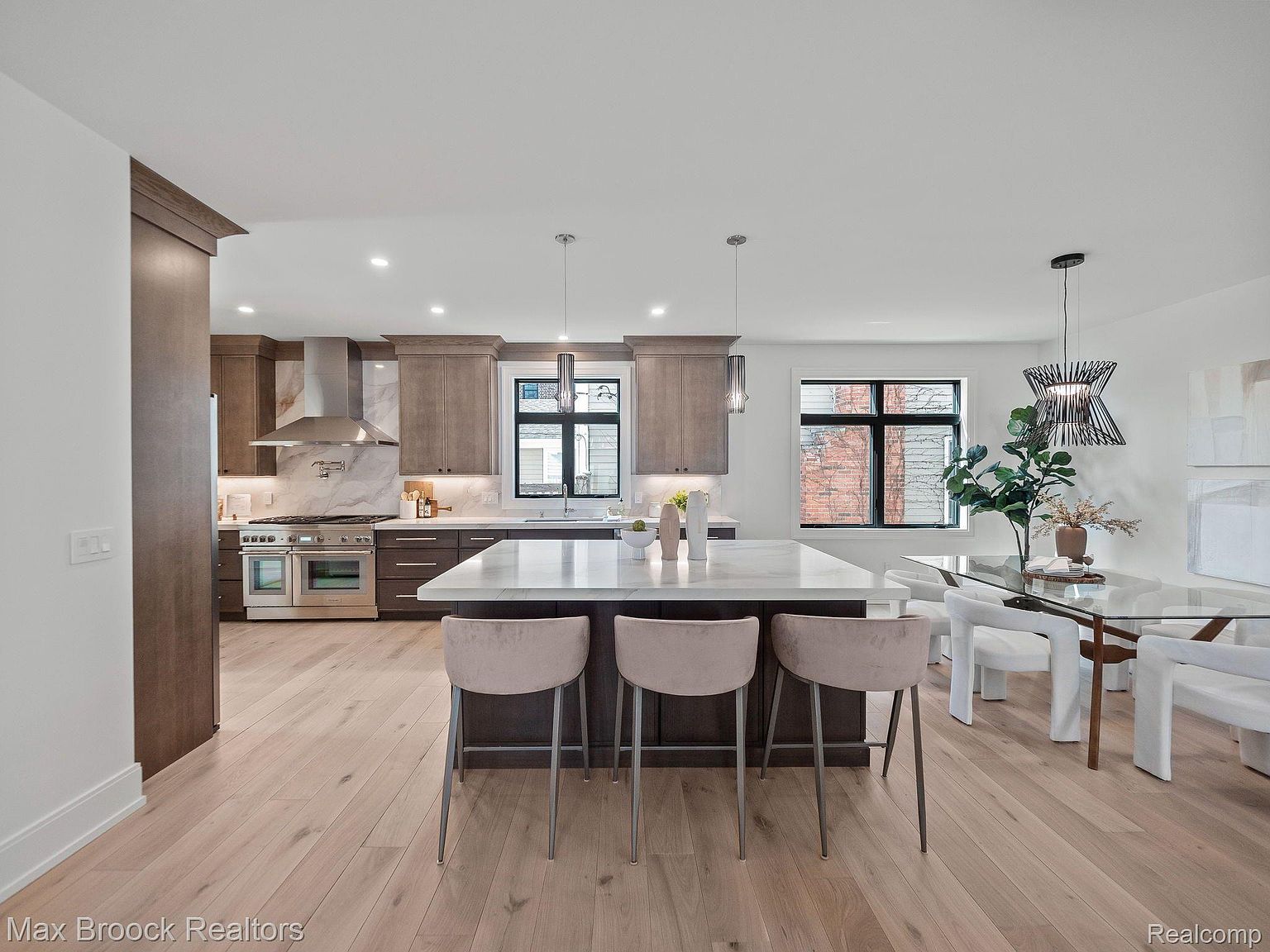 This is a bright and modern kitchen featuring a large island with seating, stainless steel appliances, and stylish pendant lighting. The kitchen has wooden cabinets and a marble backsplash, complemented by light hardwood flooring. A dining area with a glass table and unique chandelier is adjacent to the kitchen, creating an open and inviting space.
