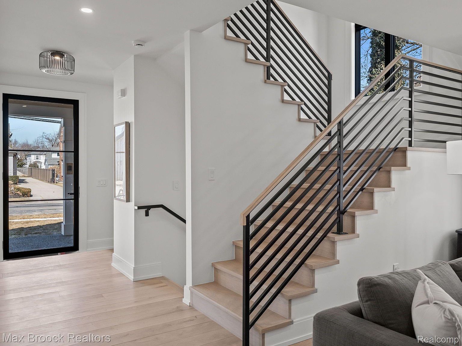 This interior shot showcases a modern hallway and staircase. The staircase features light wood treads and a striking black metal railing with horizontal bars. The hallway has light wood flooring and a black-framed glass door, contributing to a bright and contemporary aesthetic.