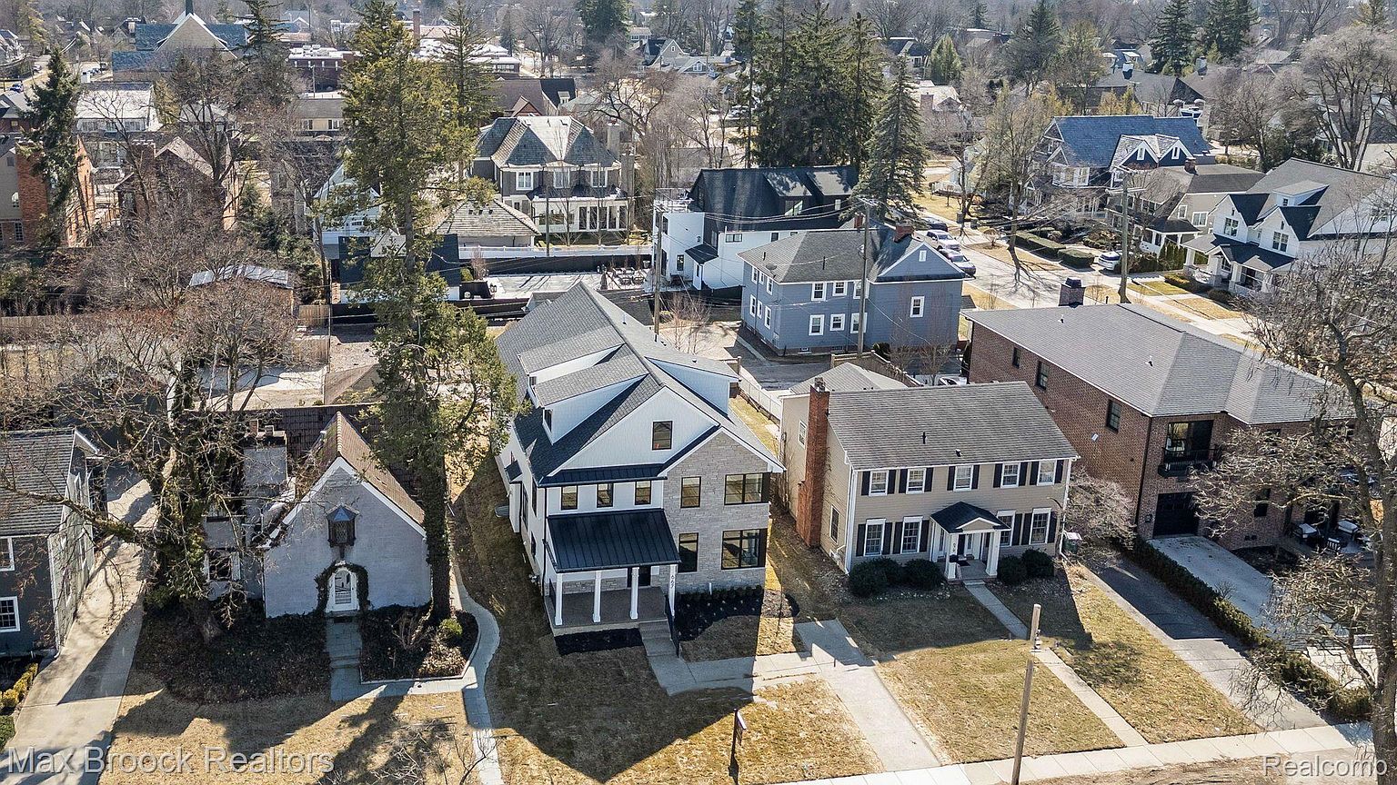 This aerial shot showcases a neighborhood with well-maintained homes, highlighting their architectural diversity and landscaping. The focal point is a modern two-story house with a gray stone facade and a black metal roof, situated between a charming cottage-style home and a classic colonial residence. The image provides a comprehensive view of the properties and their surroundings, emphasizing the desirable residential setting.