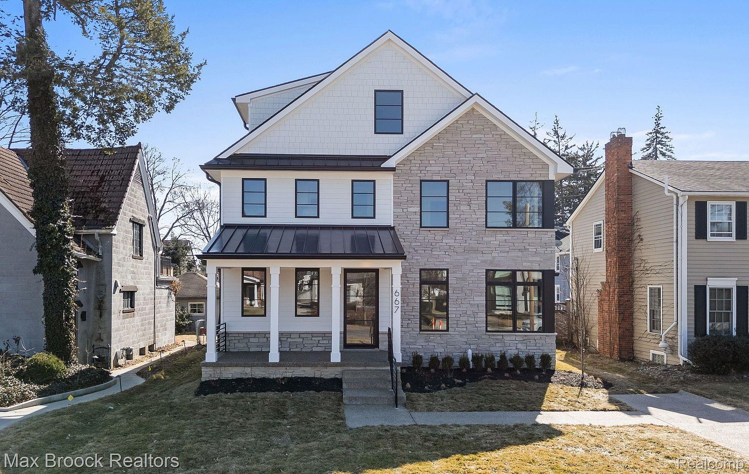 This is a front view of a two-story house with a combination of stone and white siding. The house features black-framed windows and a covered front porch with white columns. The lawn is neatly maintained, and the overall impression is one of modern elegance and curb appeal.