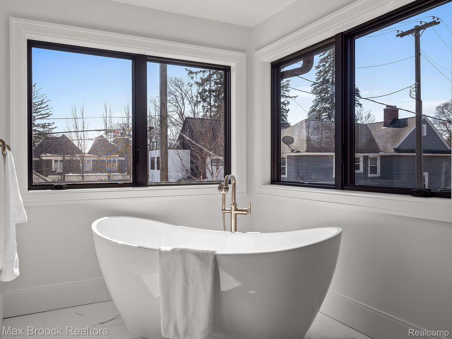 This is a bright and airy primary bathroom featuring a freestanding white bathtub positioned in front of two large windows. The windows offer a view of the neighborhood outside, bringing natural light into the space. The bathroom has a modern and minimalist design, creating a spa-like atmosphere.