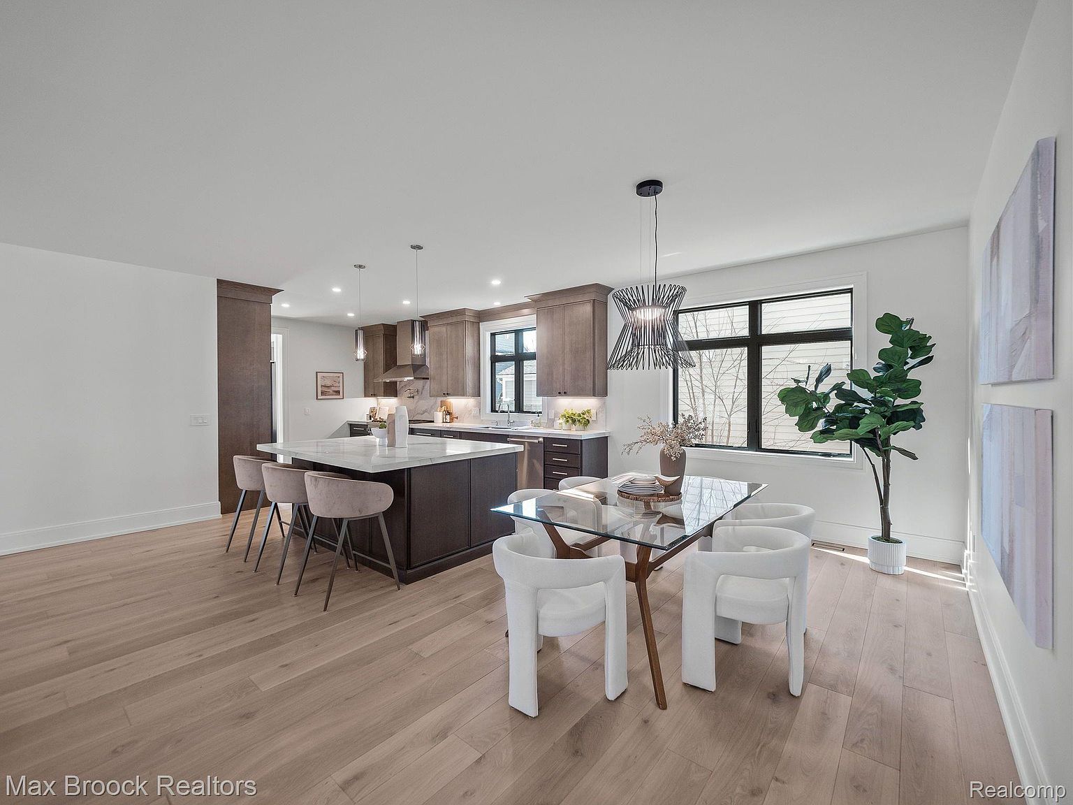 This interior shot showcases a modern kitchen and dining area. The kitchen features a large island with seating, complemented by sleek cabinetry and countertops. The dining area includes a glass-top table surrounded by contemporary white chairs, illuminated by a stylish chandelier, with a large window providing natural light and a view to the outside.