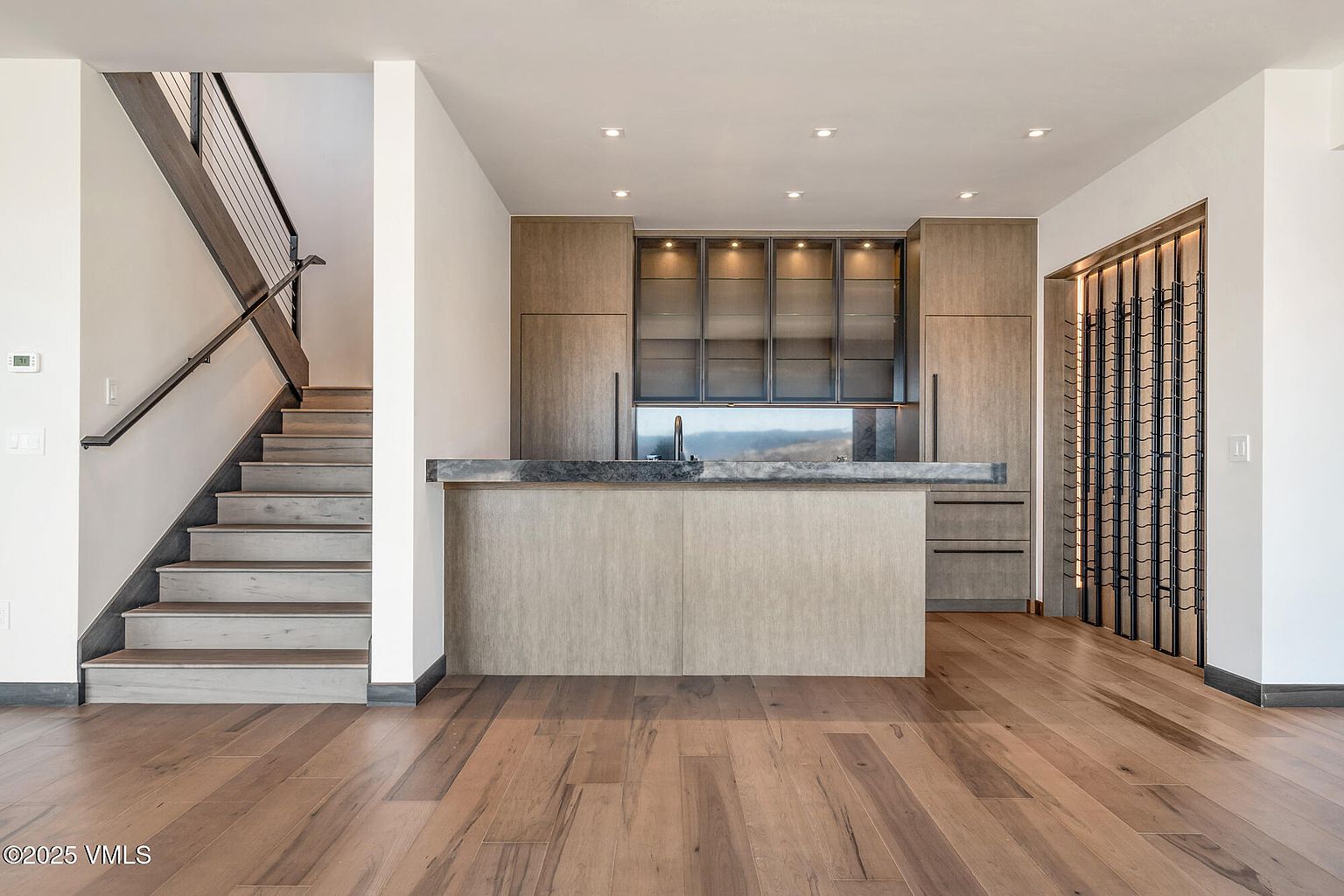 This interior shot showcases a modern kitchen with hardwood flooring and a staircase on the left. The kitchen features sleek cabinetry, a central island with a countertop, and a built-in wine rack on the right. Recessed lighting illuminates the space, creating a warm and inviting atmosphere.