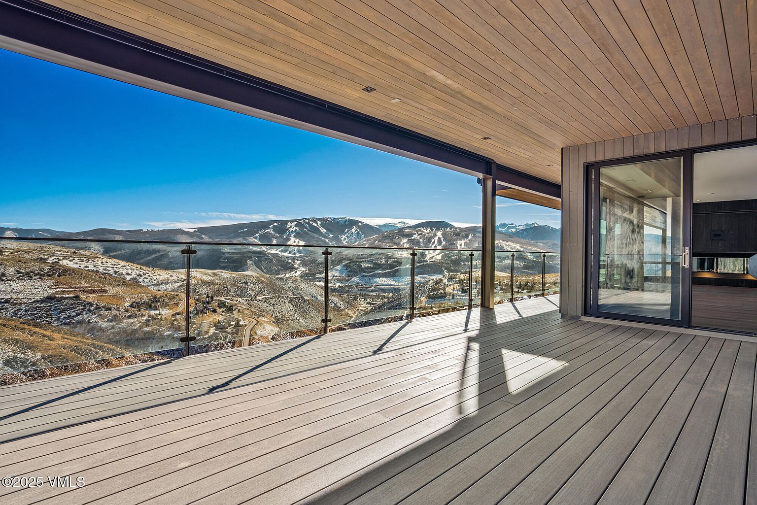 This image showcases a spacious outdoor deck with a stunning mountain view. The deck features modern design elements, including a glass railing that provides an unobstructed view of the snow-capped mountains and surrounding landscape. The wooden deck flooring and ceiling add warmth to the contemporary design, while the sliding glass doors seamlessly connect the indoor and outdoor spaces.