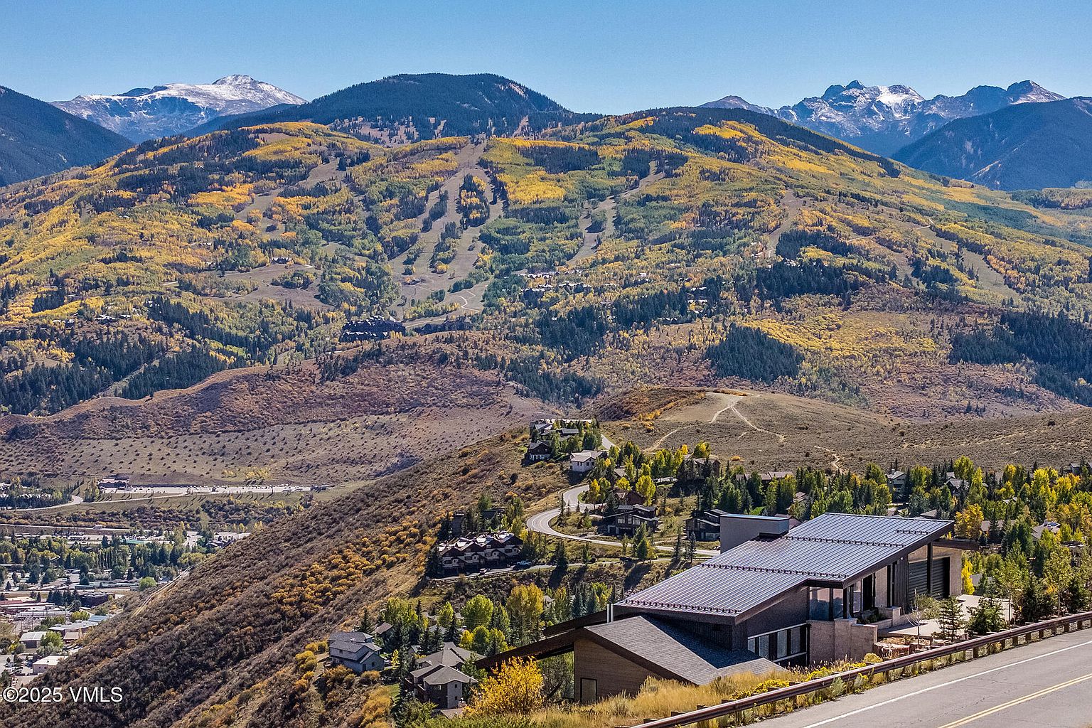 This aerial view showcases a modern home nestled on a hillside, with panoramic mountain views in the background. The house features a sleek, contemporary design with a dark roof and clean lines, blending seamlessly with the natural landscape. The surrounding area includes mature trees and winding roads, suggesting a private and upscale community.