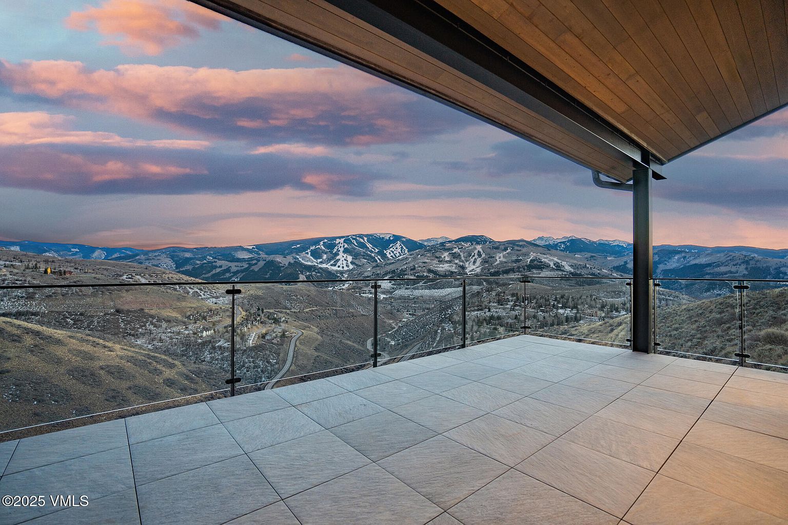 This image showcases a spacious outdoor balcony with a modern design, featuring sleek glass railings that offer unobstructed panoramic views of the surrounding mountains and landscape. The tiled flooring adds a touch of elegance, while the wooden ceiling provides a warm contrast. The scene is bathed in the soft light of dusk, creating a serene and inviting atmosphere.