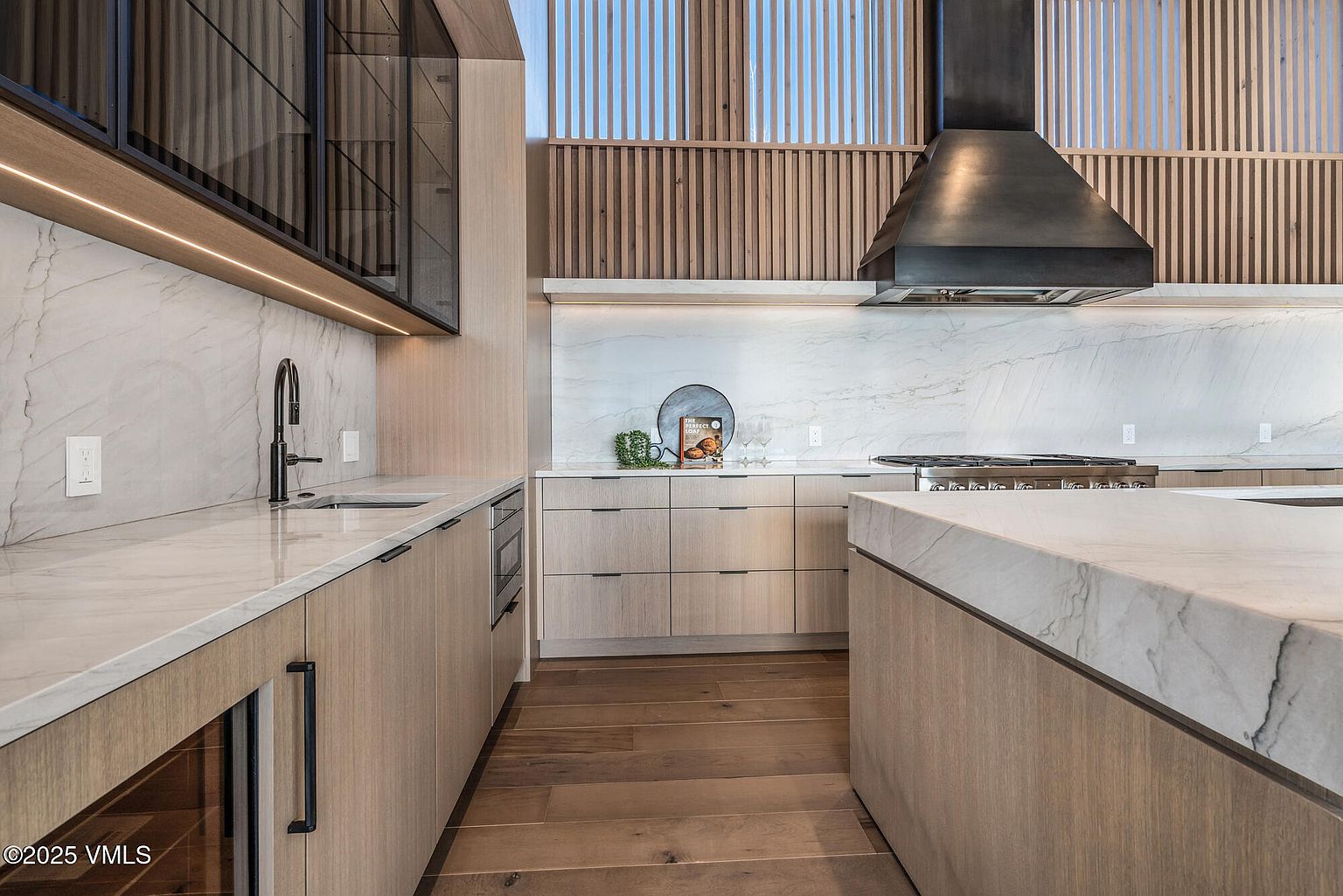 This is a modern kitchen featuring light wood cabinetry, marble countertops and backsplash, and stainless steel appliances. A large island provides ample counter space, and a black range hood adds a touch of contrast. The perspective is from the entrance of the kitchen, showcasing the layout and design elements.